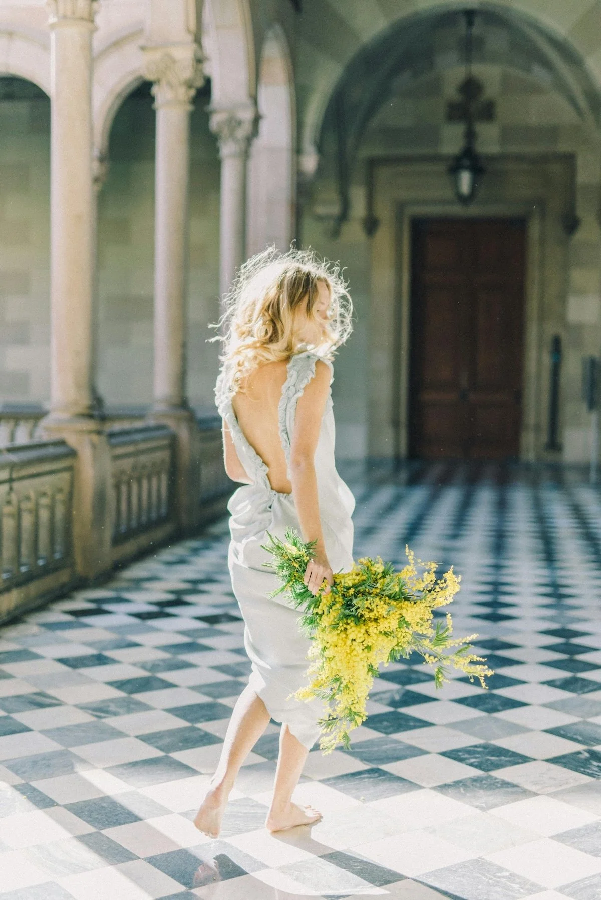 A woman in a backless white dress holding a large bouquet of yellow flowers, standing barefoot in a sunlit, ornate hallway with checkered black and white floor tiles and arched columns.
