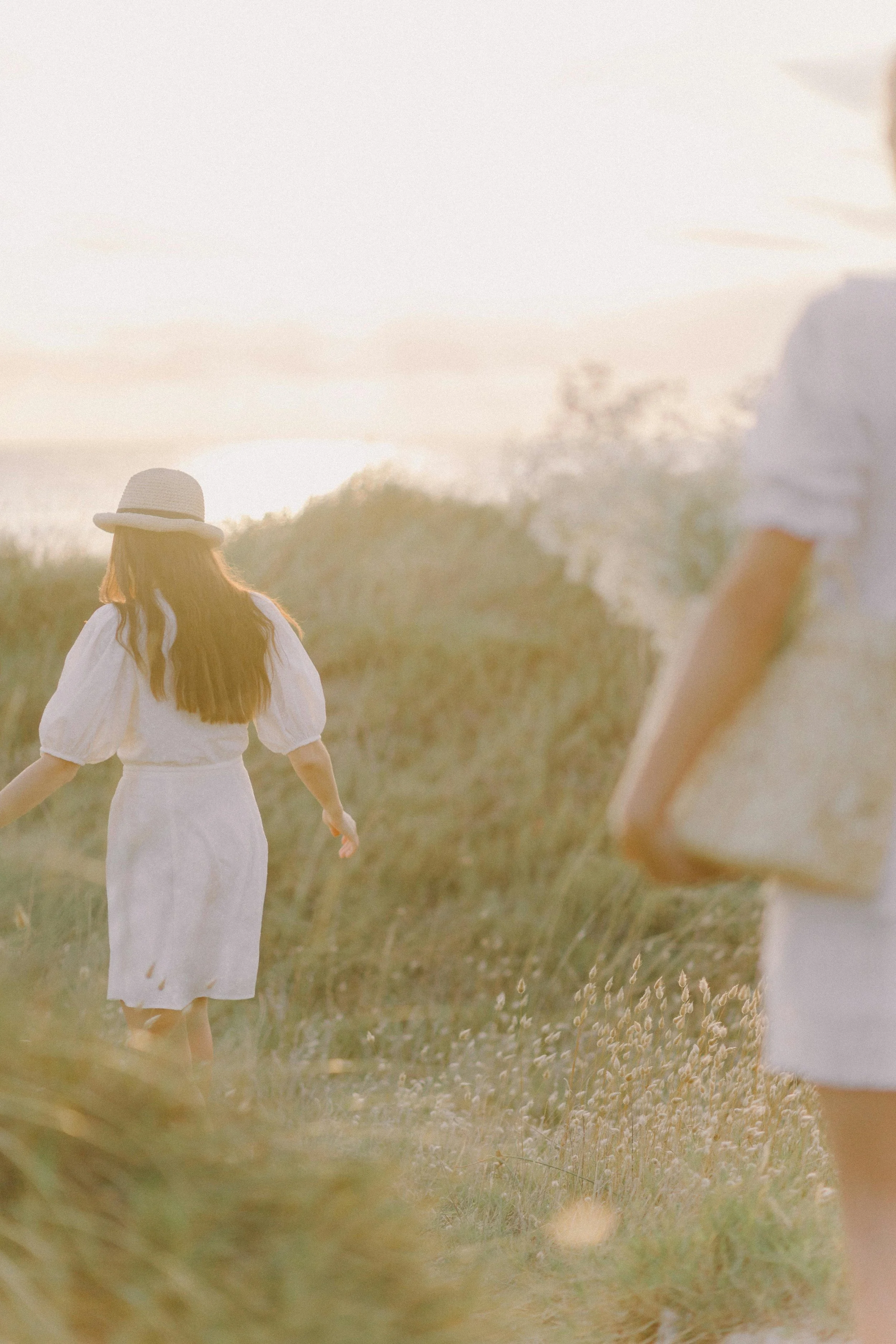 Two women in a field with tall grass during sunset. One woman wears a white dress and sunhat, facing away from the camera. The other woman, partially visible, wears a white top with rolled sleeves.