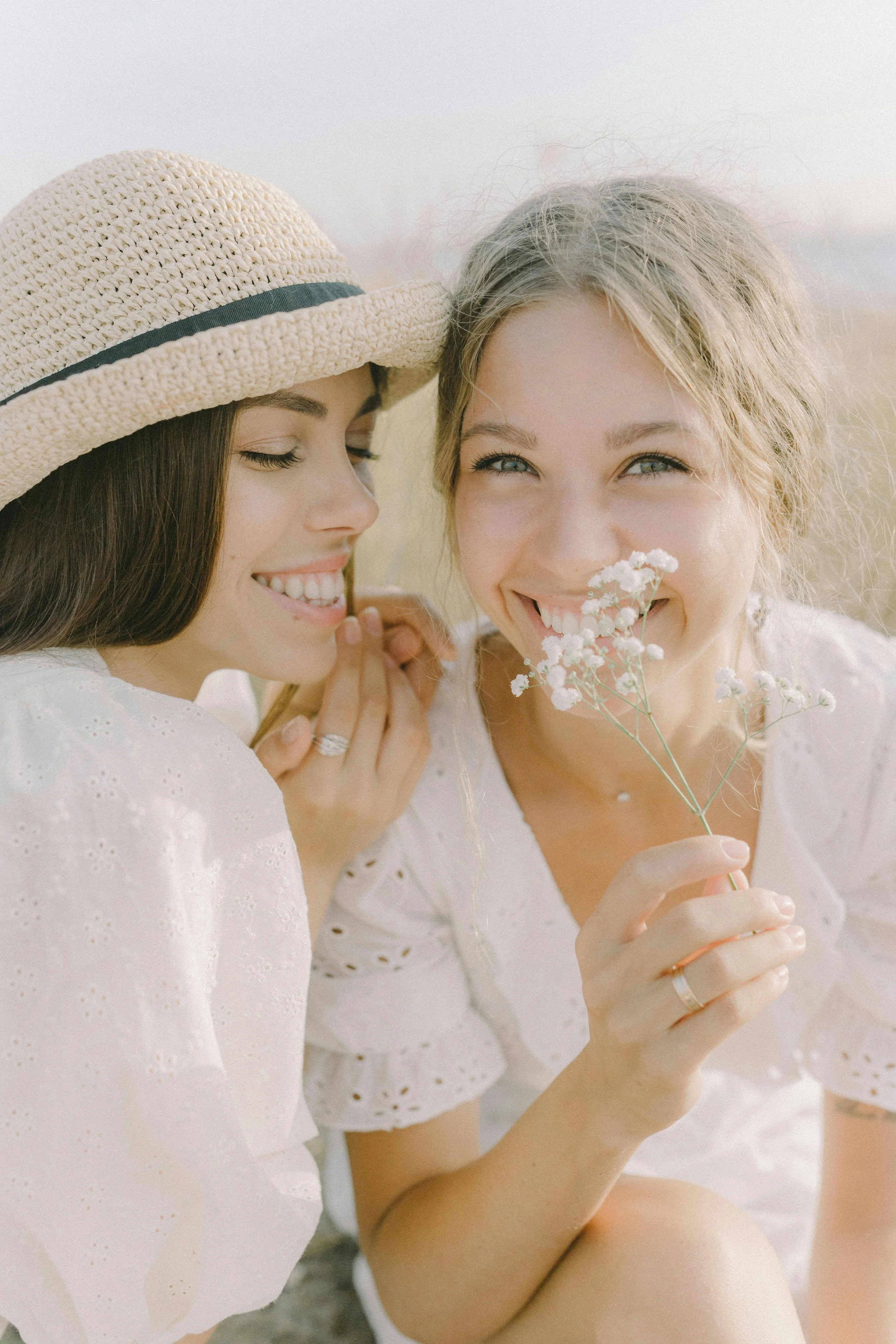 Two women smiling and holding a small bouquet of white flowers outdoors during daytime. One woman wears a straw hat and the other has light blonde hair and blue eyes. Both wear white clothing with eyelet details.