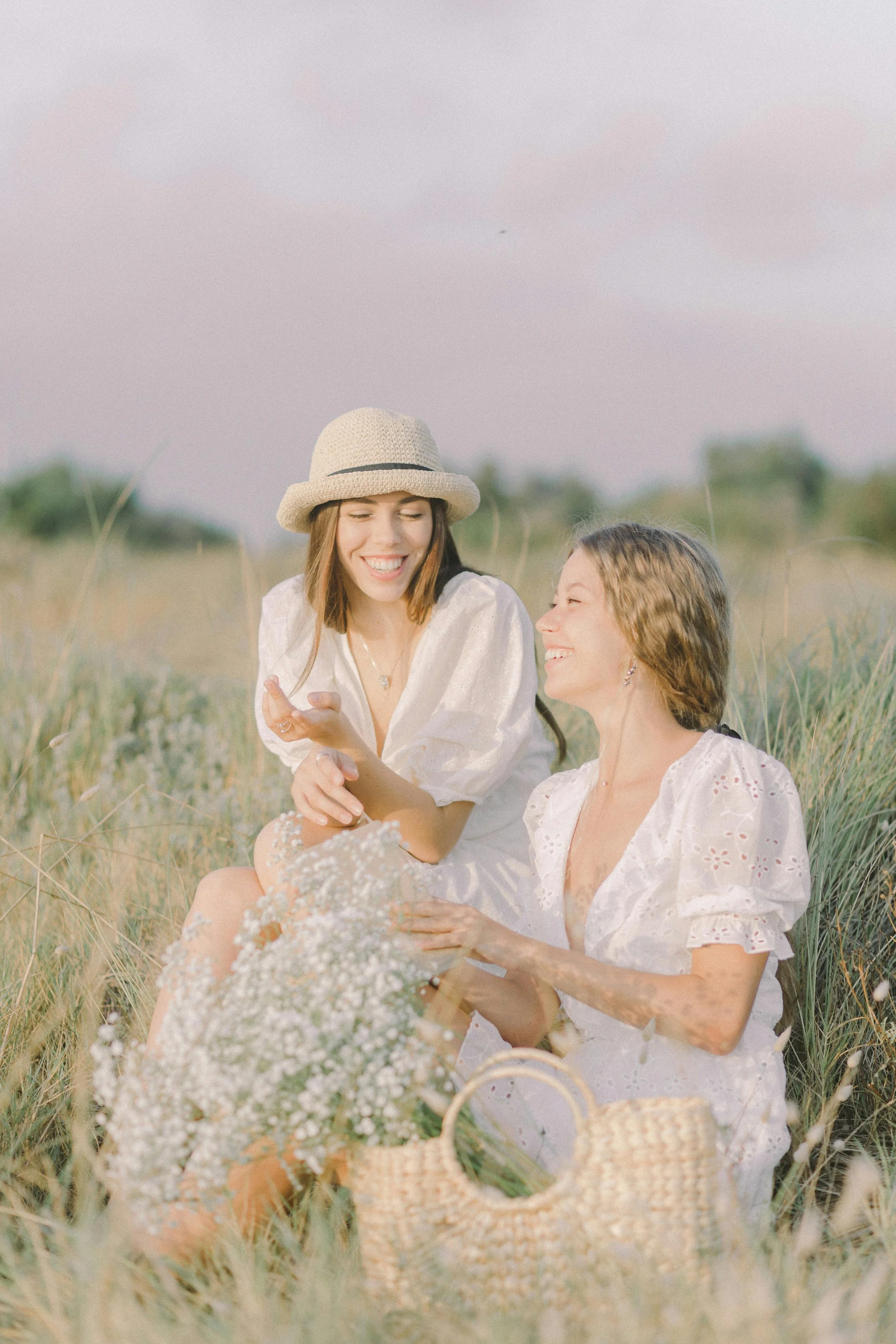 Two women sitting in a field of tall grass with flowers, smiling and interacting, wearing white dresses and one has a straw hat.