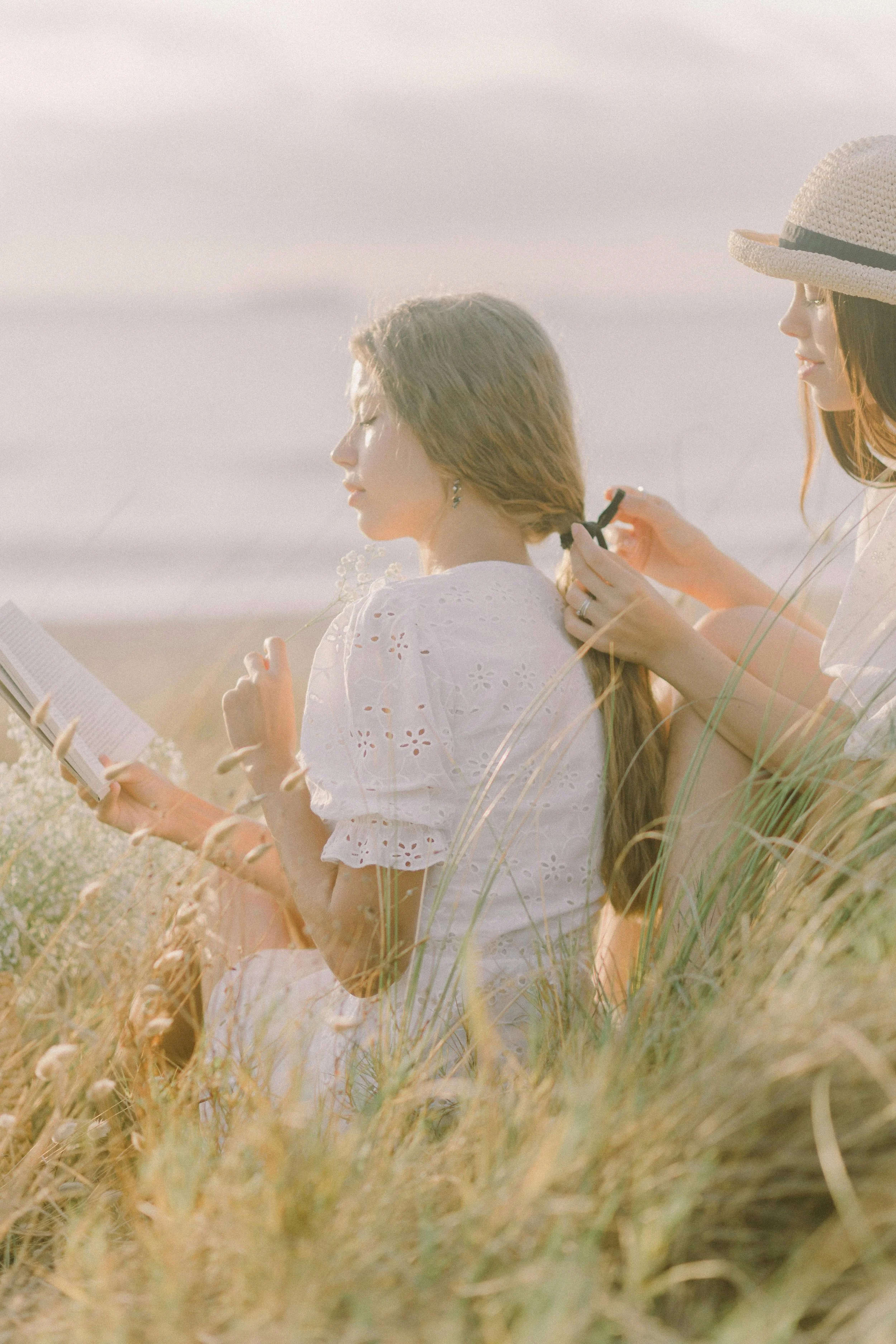 Two young women sitting in tall grass on a sunny day, one reading a book and the other braiding her hair, both dressed in light, summery clothing.