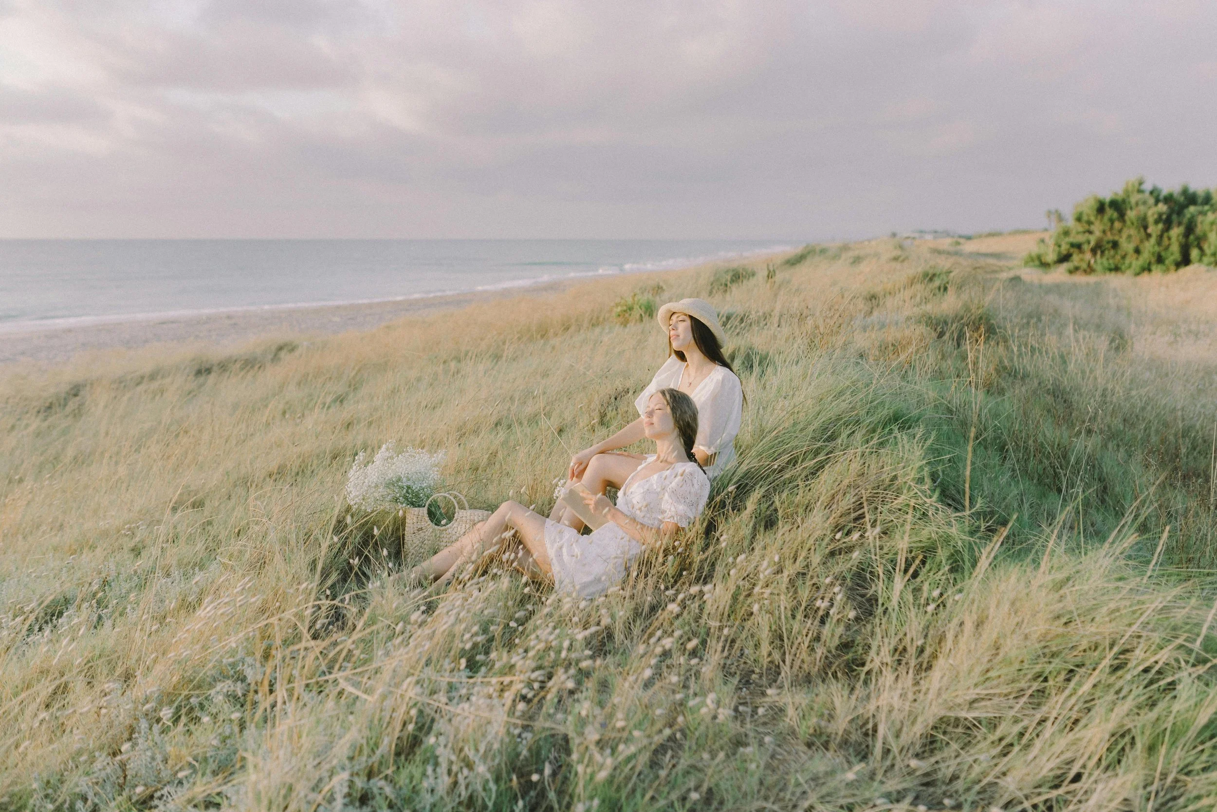 A woman and a girl relaxing in a grassy dunes near the beach, with cloudy skies and the ocean in the background. The woman wears a sun hat and a white dress, while the girl holds a book.