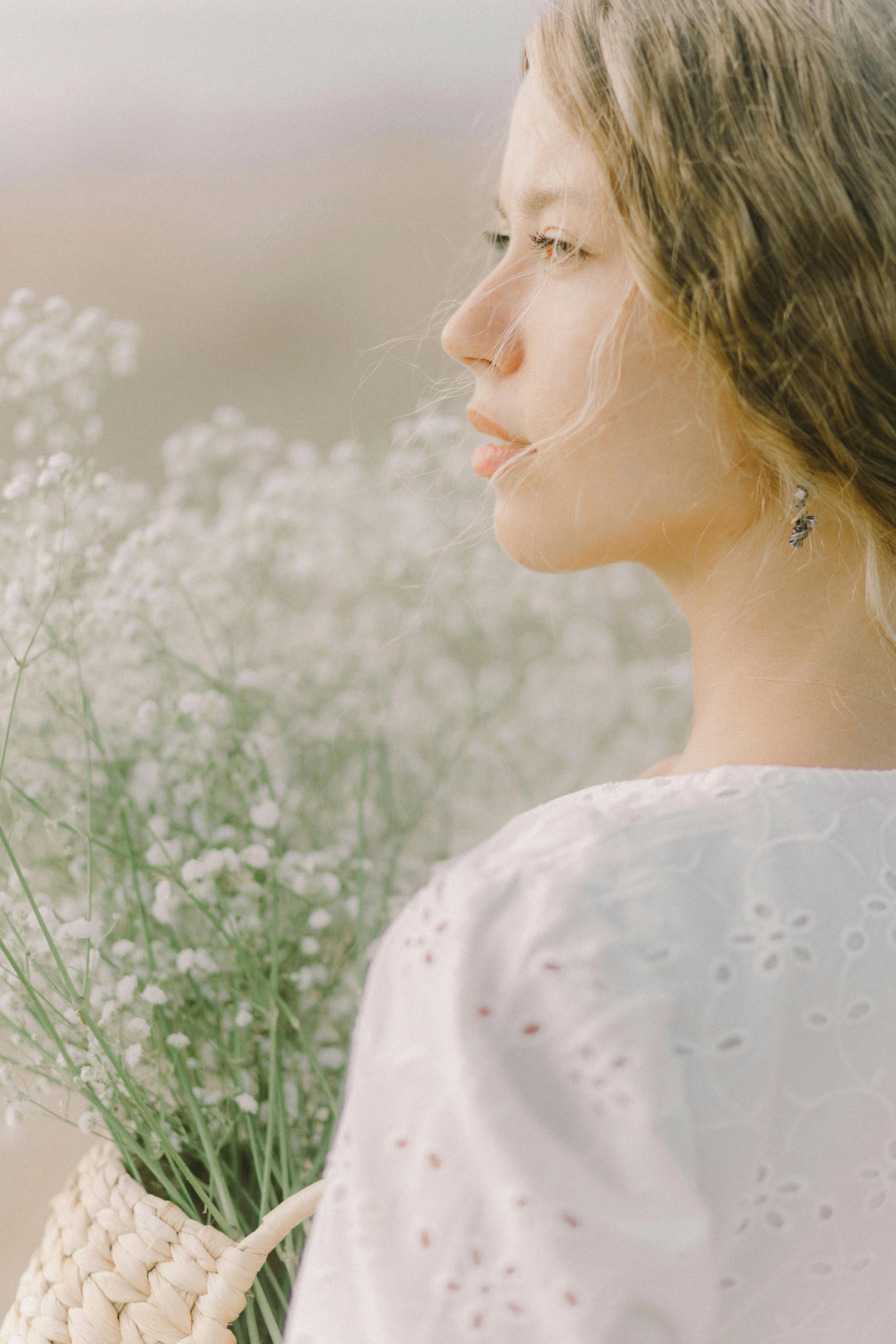 A young woman with blonde hair and earrings holds a basket of white flowers, looking to the side in a soft, natural setting.