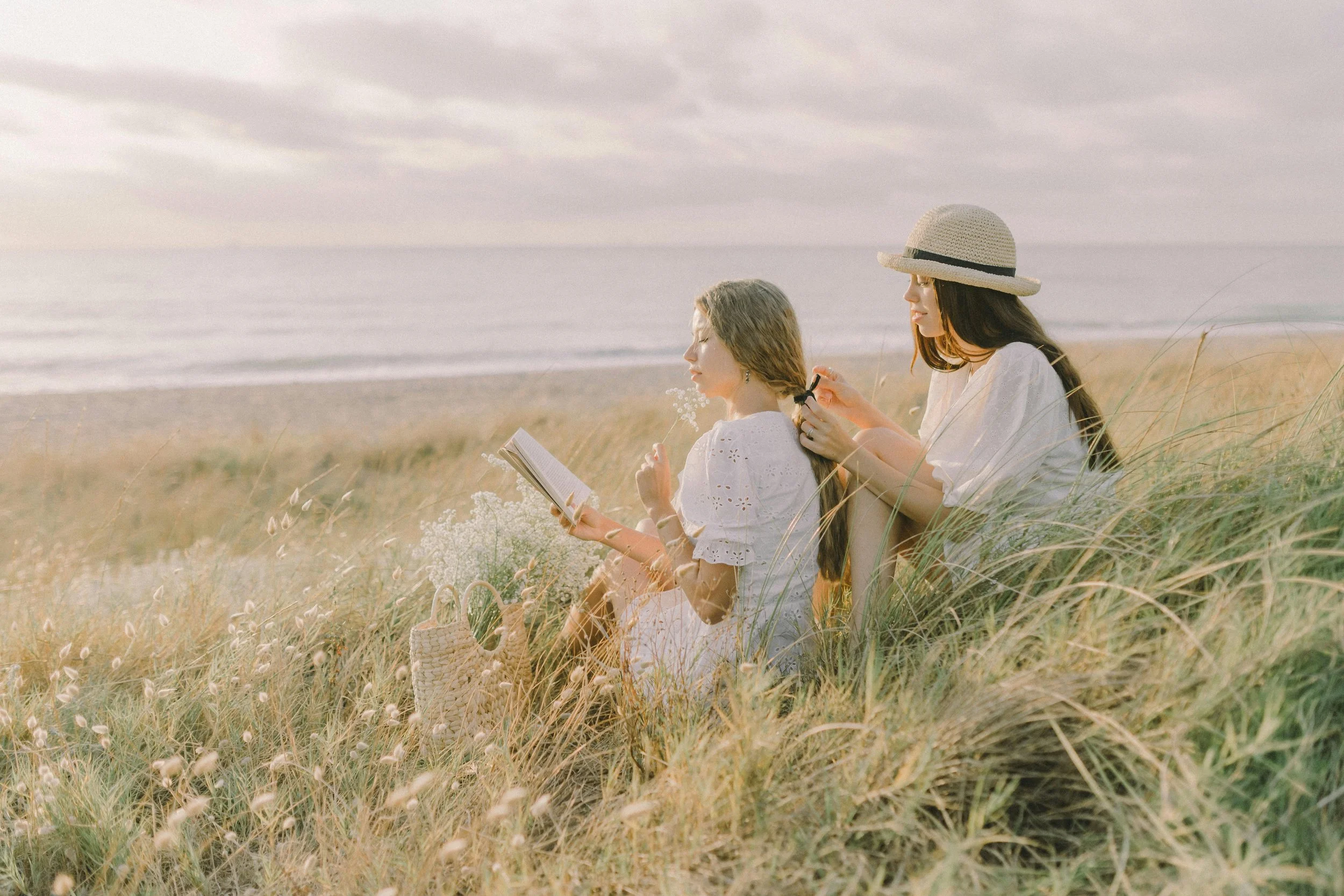 Two women sit on a grassy beach at sunset, one reading a book and the other braiding her hair, with the ocean and cloudy sky in the background.