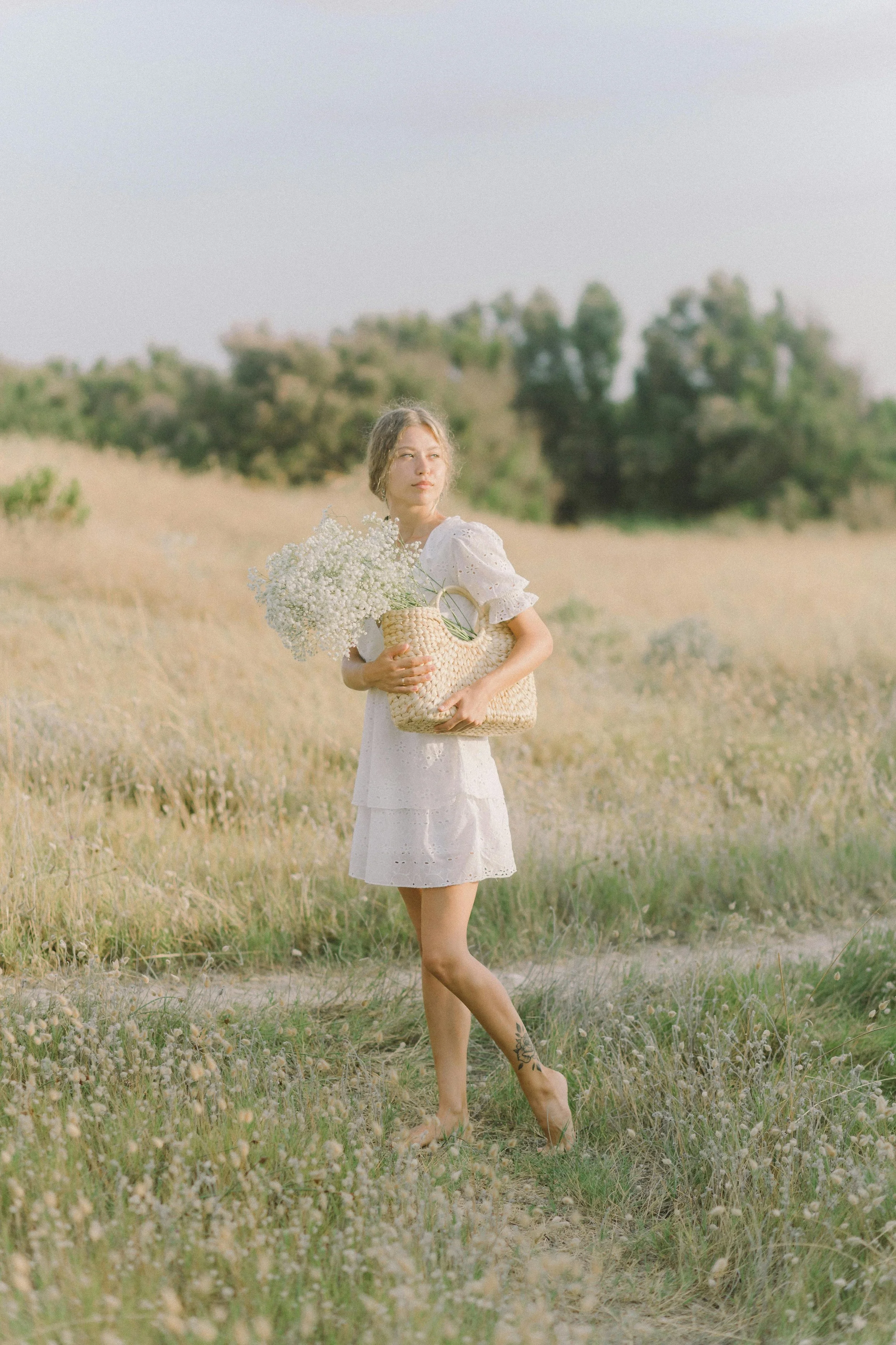 A young woman stands barefoot in a field of tall grass, holding a large wicker basket filled with white wildflowers, wearing a white dress with puffed sleeves, gazing into the distance with trees in the background under a clear sky.