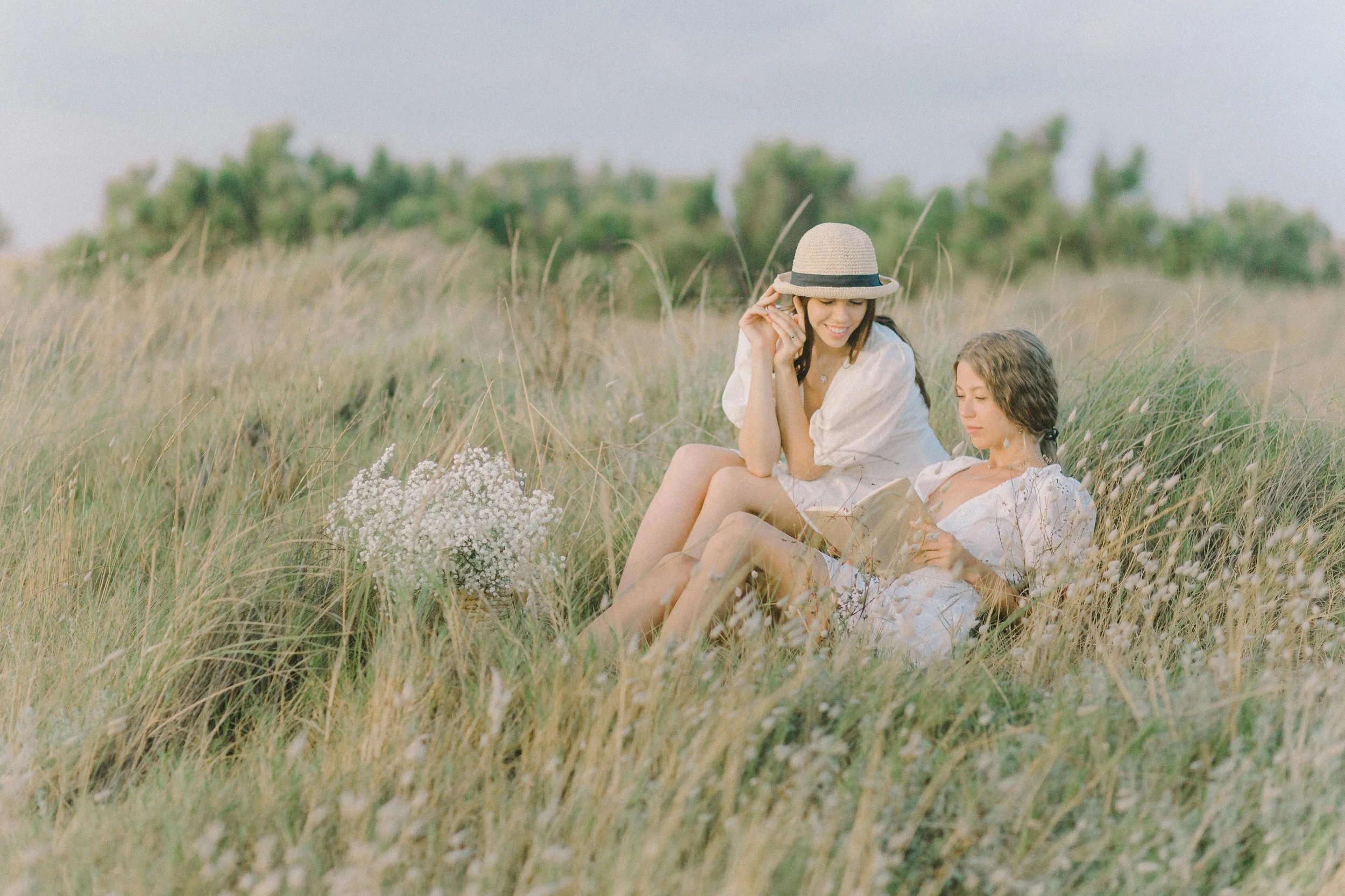 Two women sitting in a grassy field with wildflowers, one wearing a straw hat and the other reading a book.