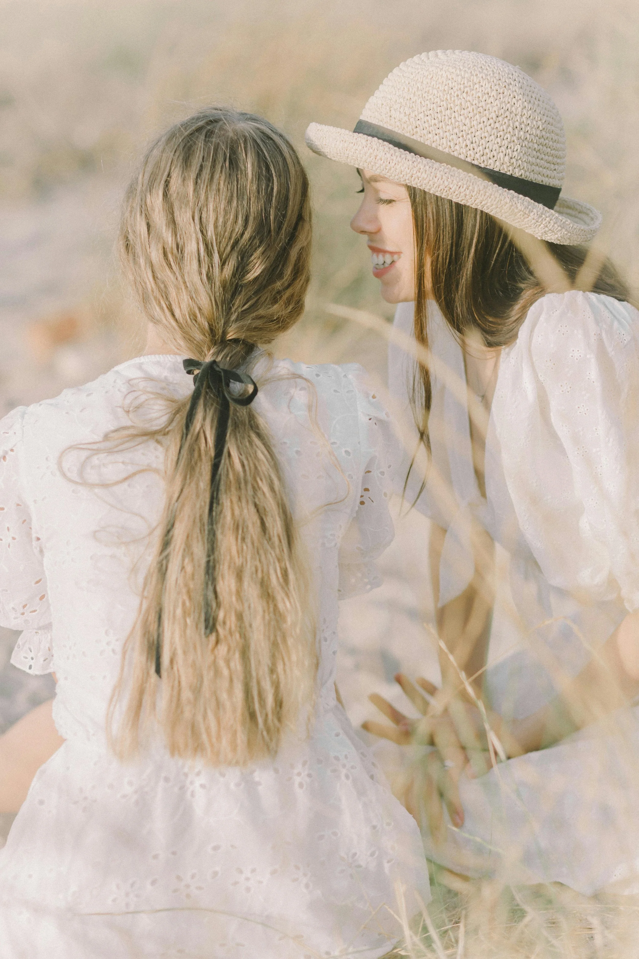 A woman wearing a wide-brimmed straw hat and white dress with eyelet details, leaning with a girl with long wavy hair tied with a black ribbon, in a natural outdoor setting.