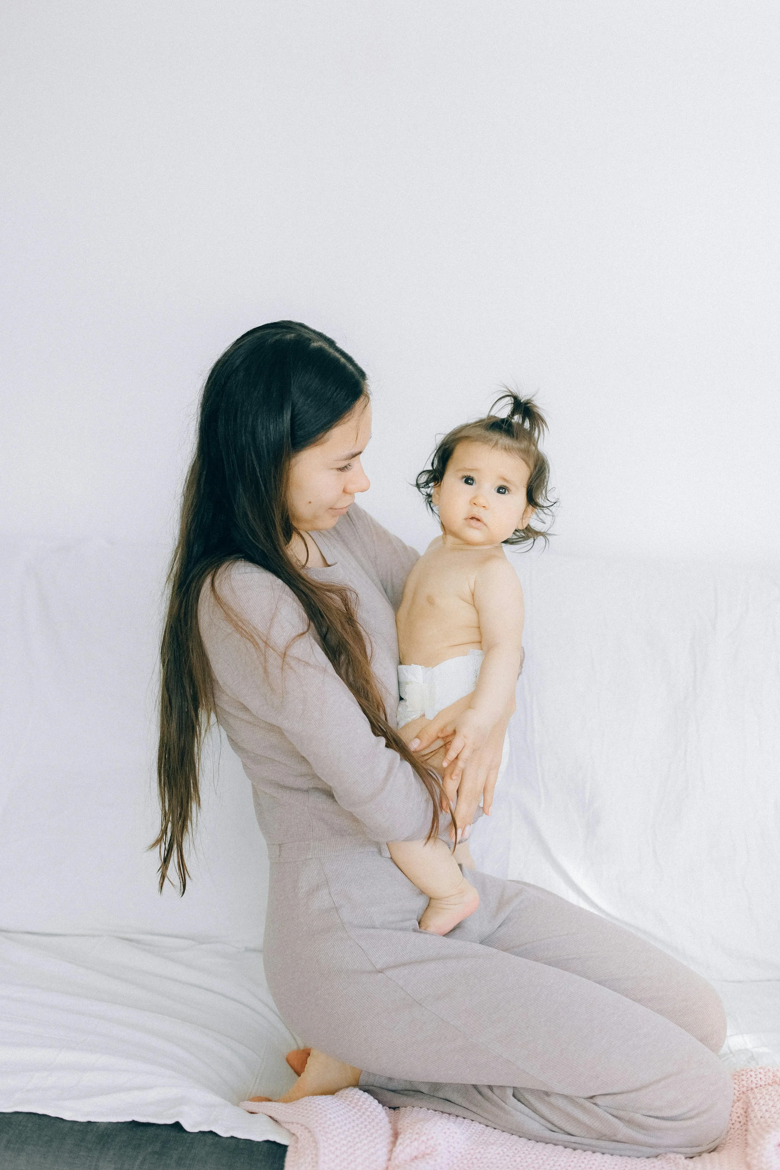 A woman with long dark hair kneeling on a bed, holding a young toddler with curly hair and a puzzled expression, against a plain white background.