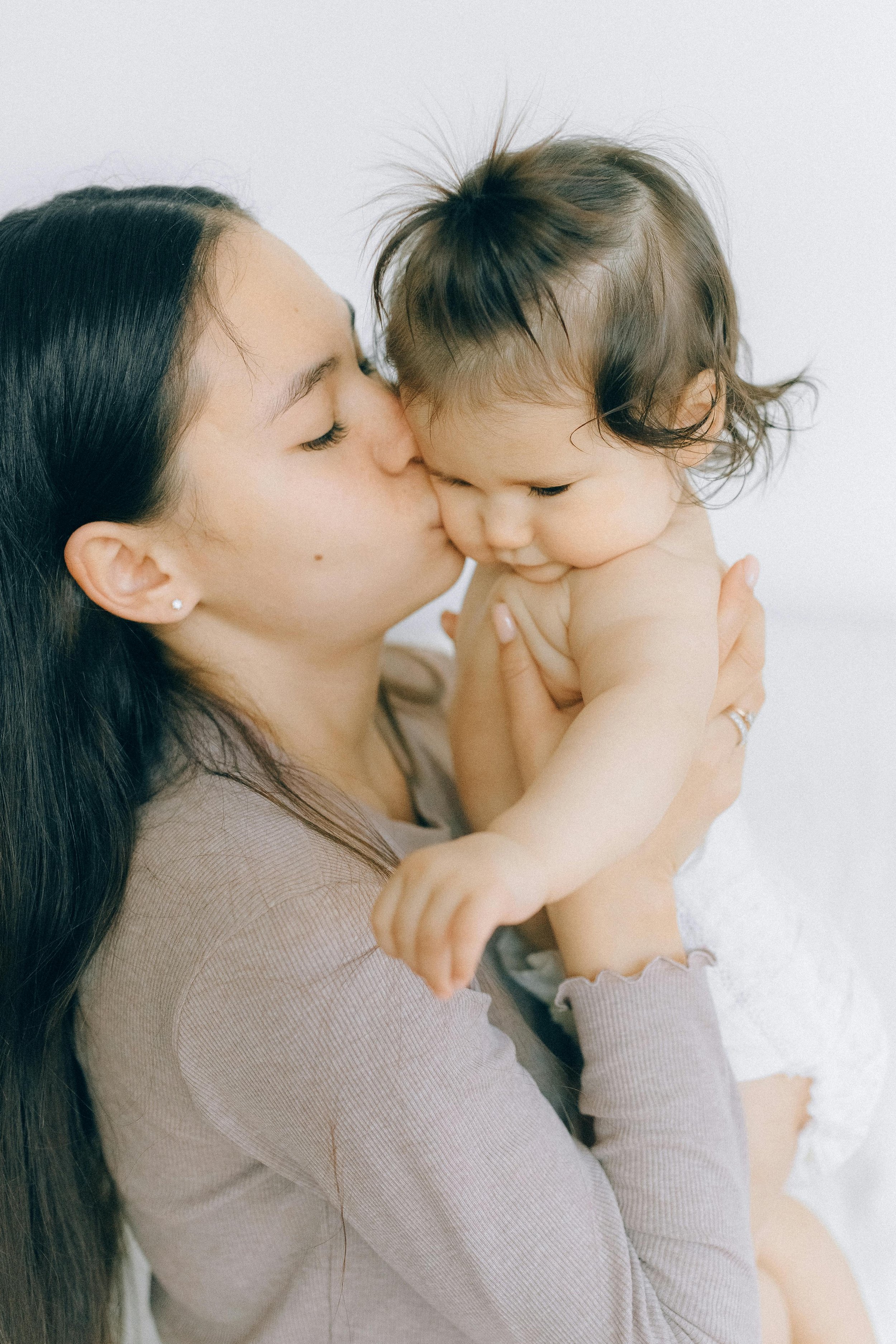 A woman with long dark hair and pierced ears gently kisses a young child with tousled dark hair on the forehead, both in close embrace against a plain white background.