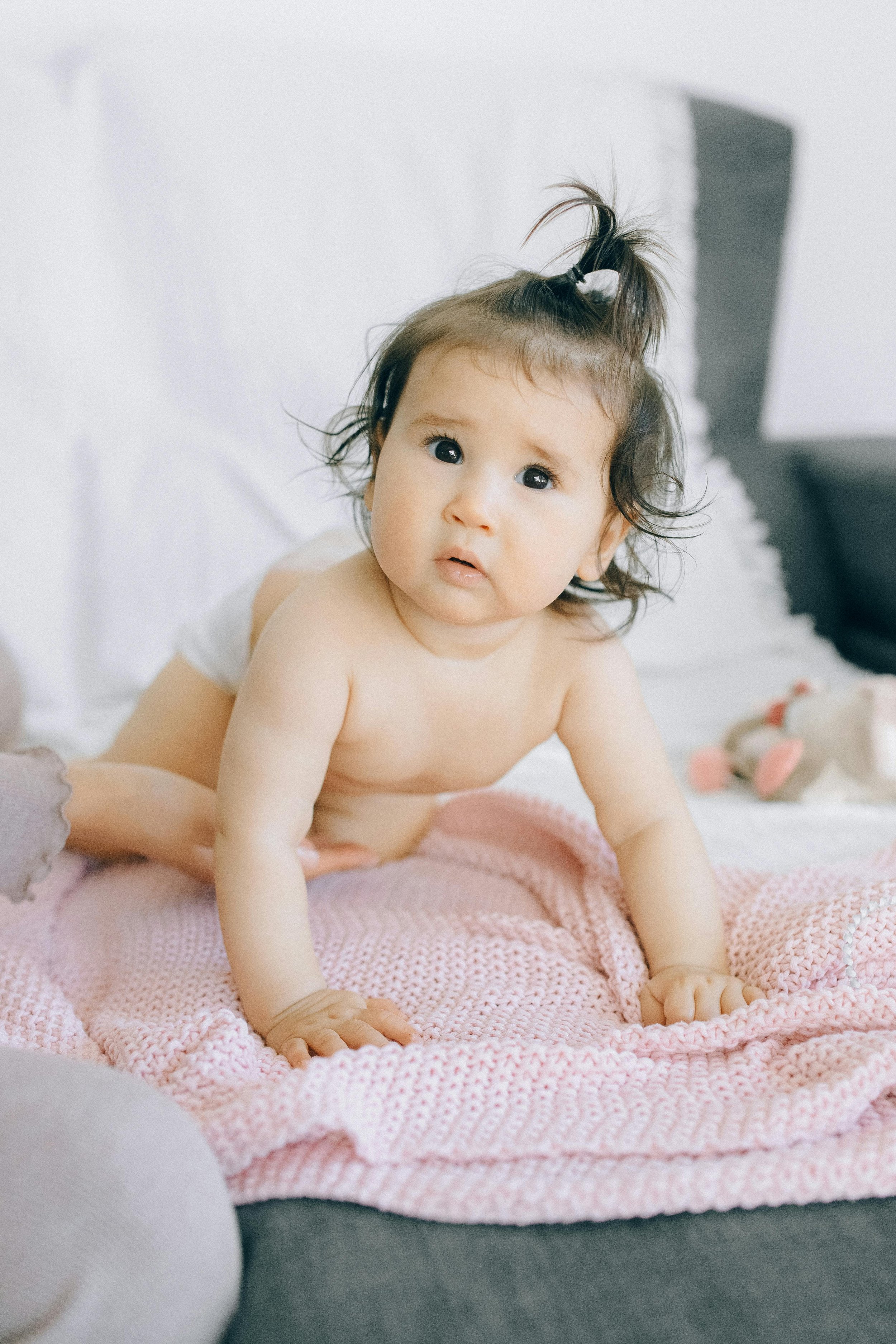 A baby with dark hair styled in a high ponytail, lying on a pink knitted blanket on a bed, looking up with a curious expression. There is a stuffed animal visible in the background.