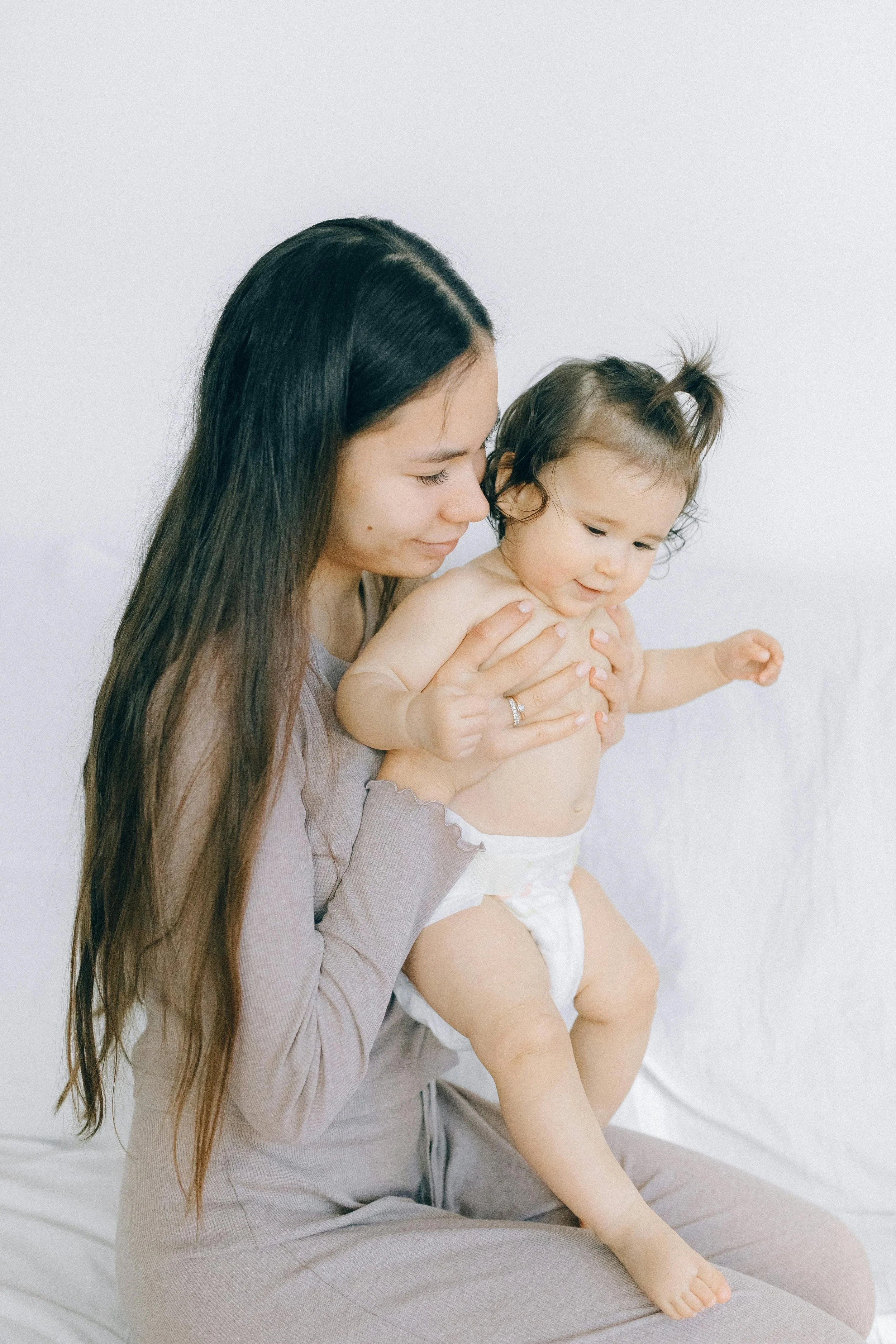A woman with long dark hair holding a smiling baby girl with a high ponytail on her shoulder against a plain light background.