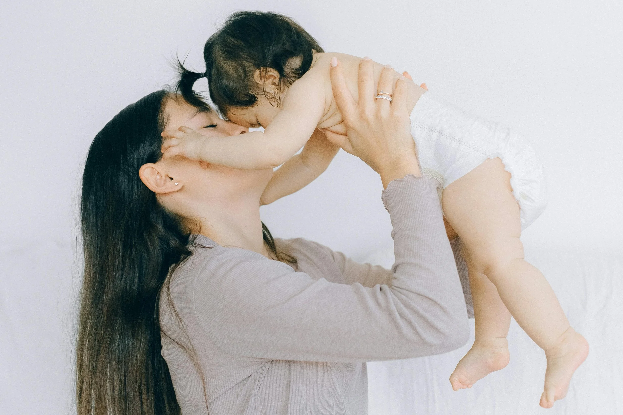 A woman holding a young girl in the air, with their faces close and eyes closed, sharing a tender moment, against a plain white background.
