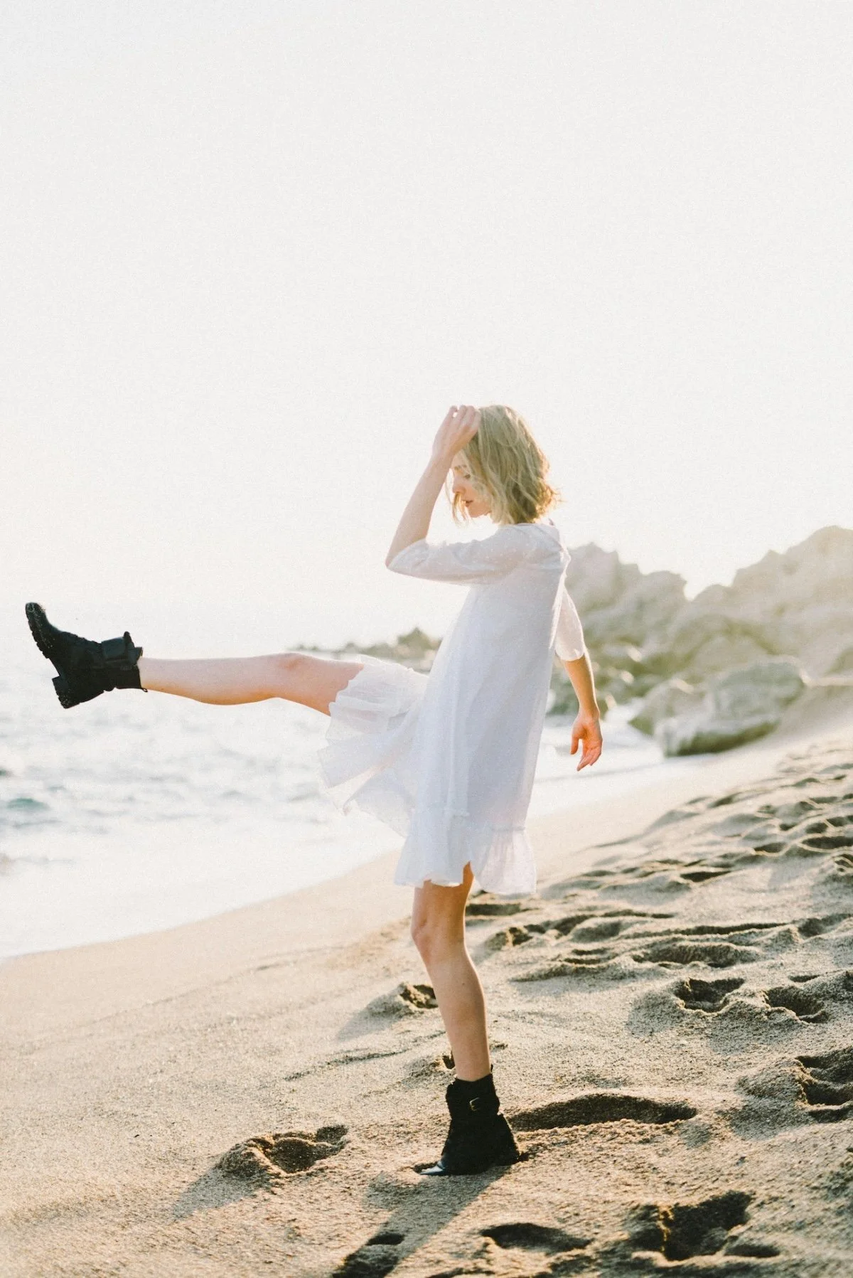 A young woman in a white dress and black boots playing on a sandy beach, kicking her leg up, with rocks in the background and the sun shining brightly.