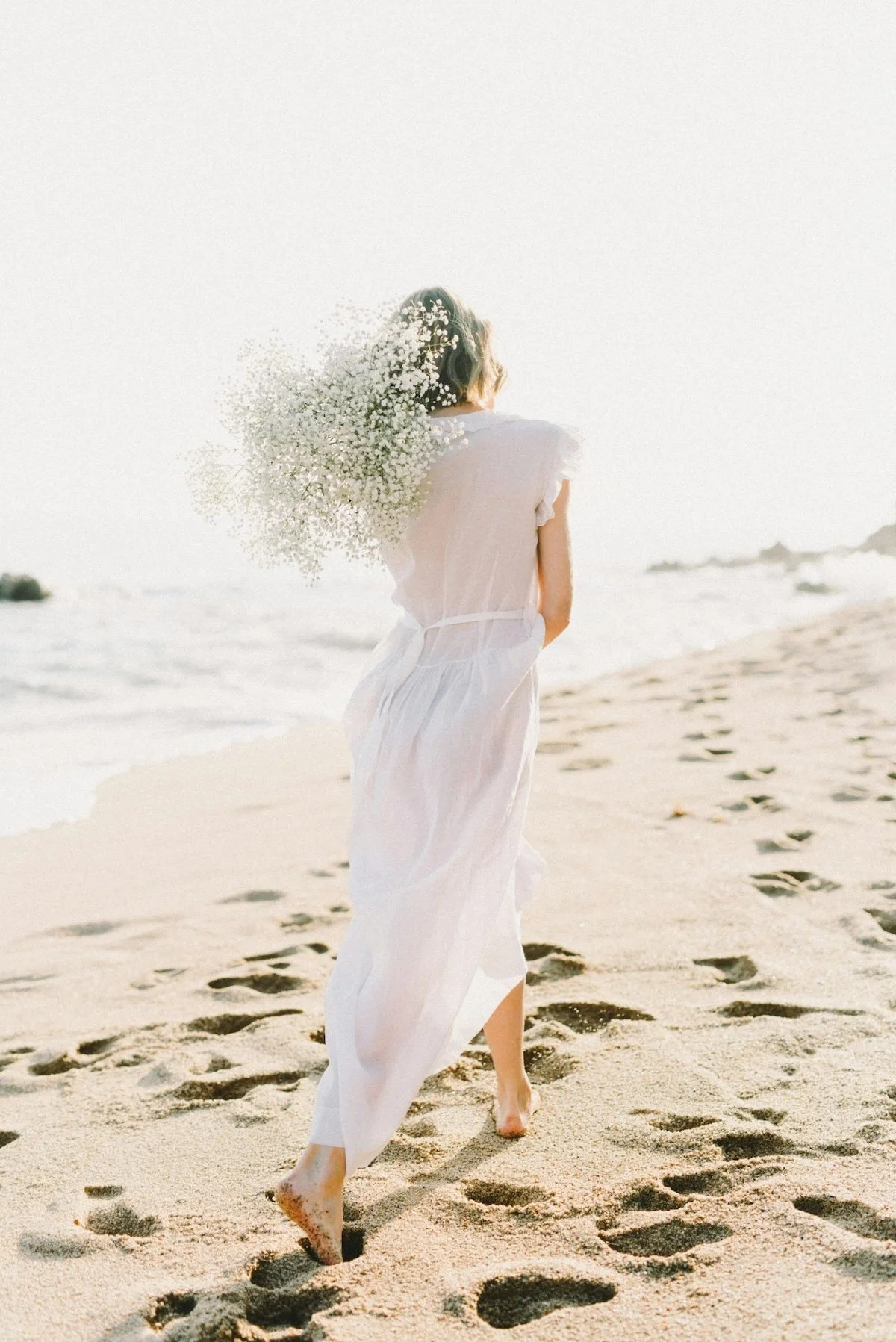 Woman in a white dress walking on the sandy beach near the shoreline during daytime, with the ocean and distant rocks in the background.
