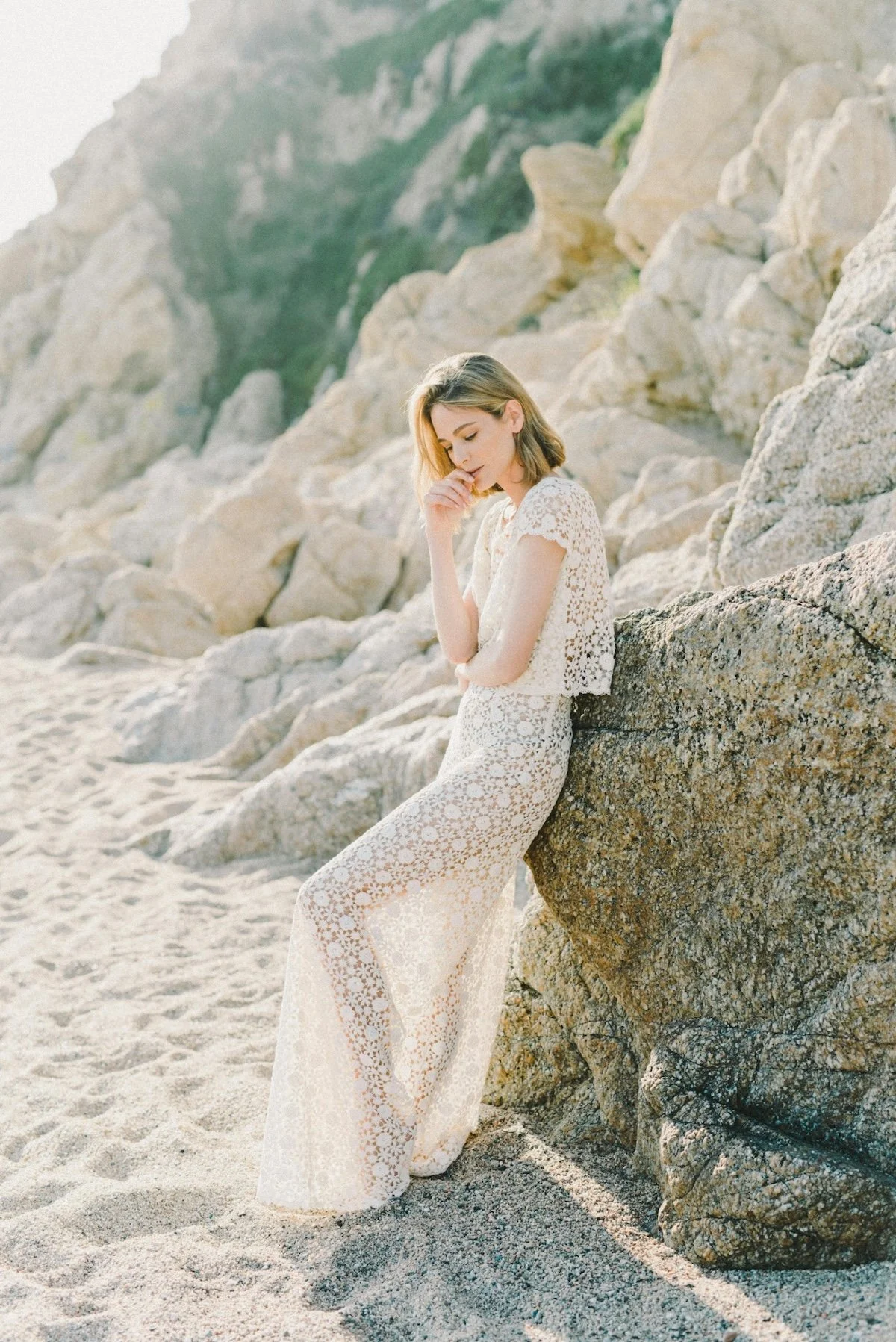 A woman in a white lace outfit leaning against a large rock on a sandy beach with rocky cliffs in the background.