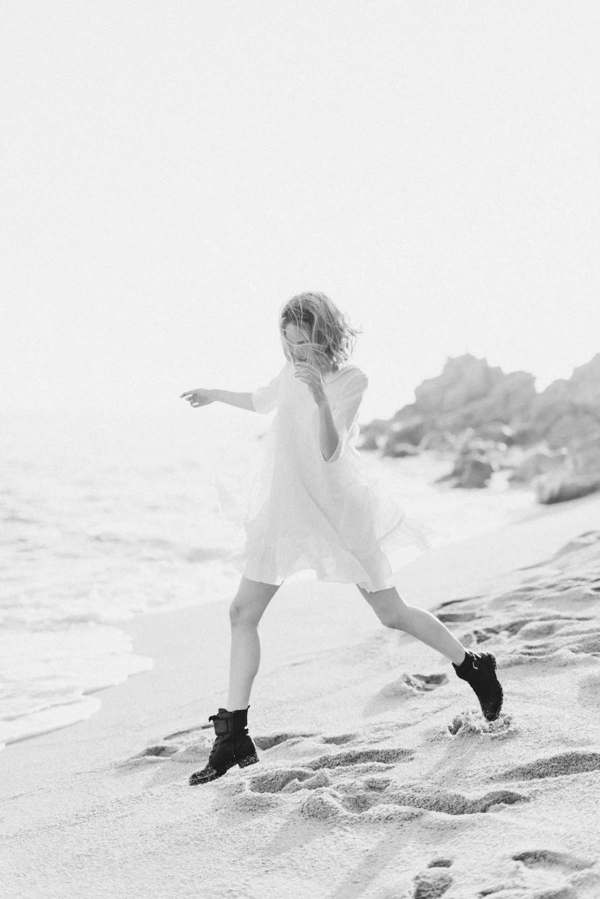 A woman in a dress and boots is jumping and playing on the beach near rocks with the ocean in the background.