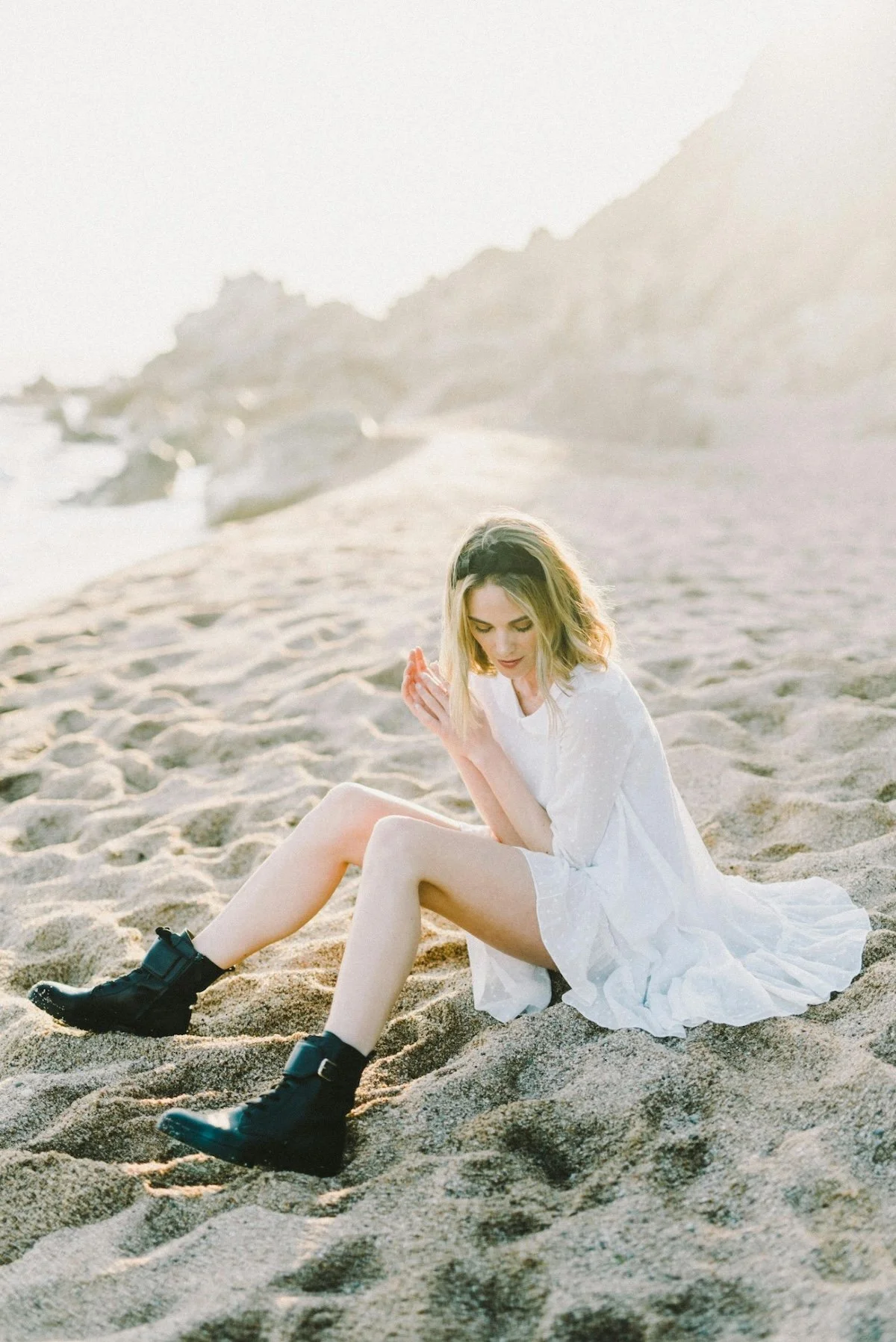 A young woman with blonde hair sitting on a sandy beach, wearing a white dress and black boots, with rocky cliffs in the background during sunset.