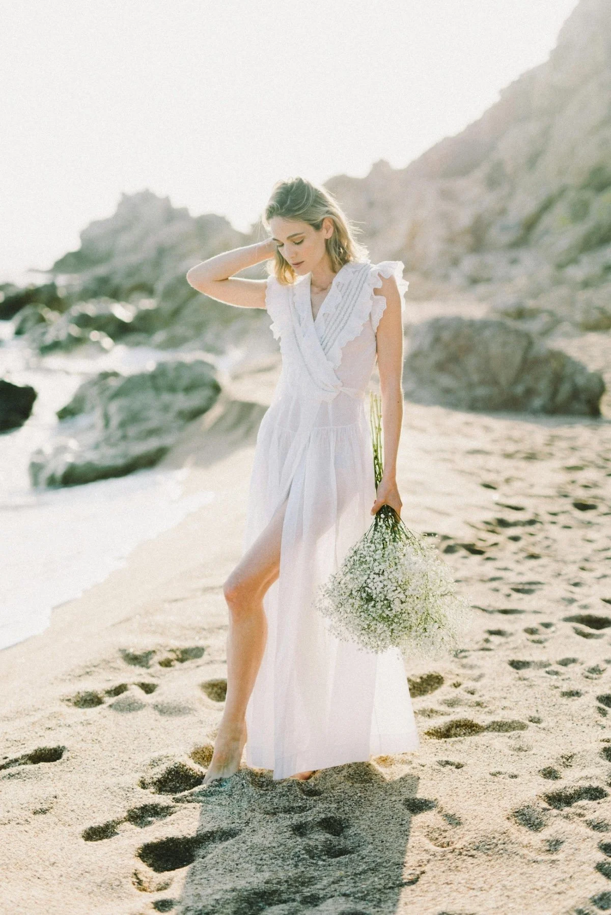 A woman in a white dress holding a bouquet of small white flowers walking barefoot on a sandy beach with rocks and hills in the background.