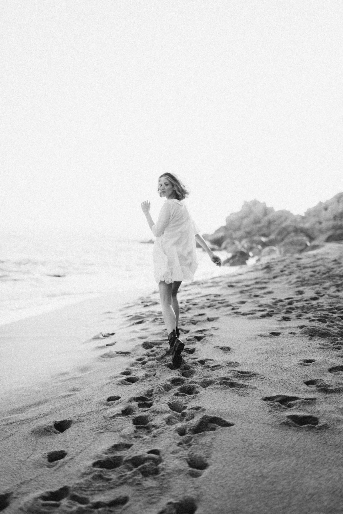 A young woman walking on a sandy beach, wearing a light-colored dress and boots, with footprints in the sand behind her and rocks in the background.