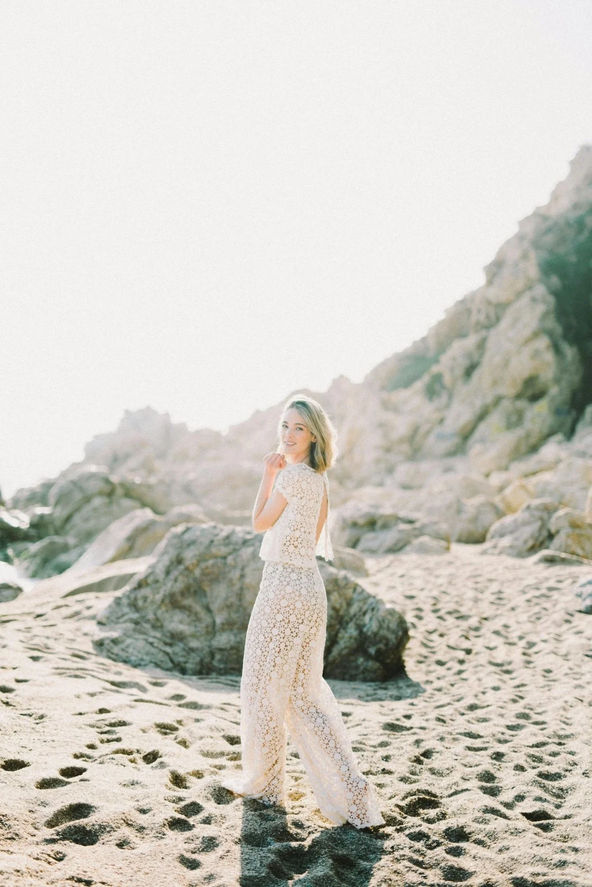 A woman in a white lace top and matching lace pants stands on a sandy beach with rocks and cliffs in the background, smiling at the camera.