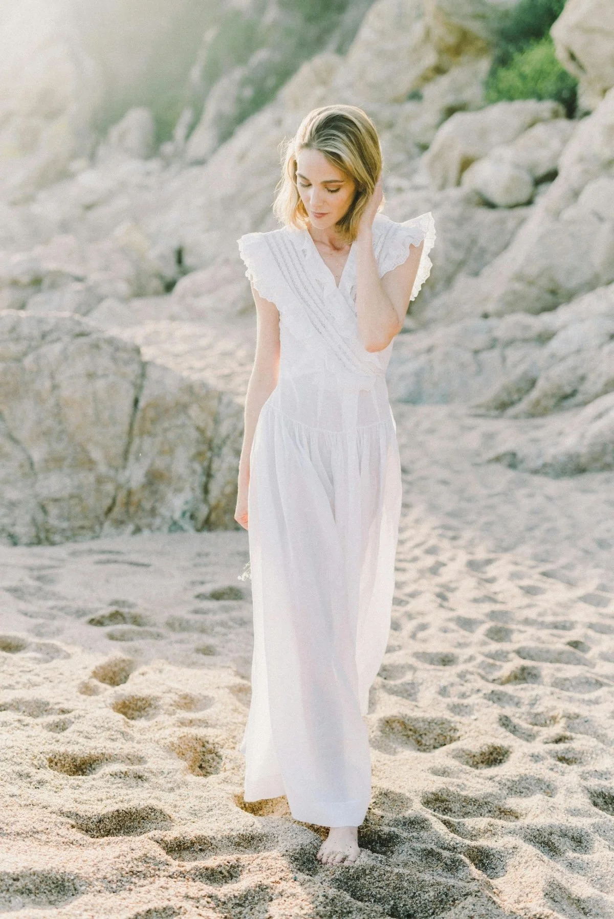 A woman in a white dress standing barefoot on a sandy beach with rocks in the background during sunset.