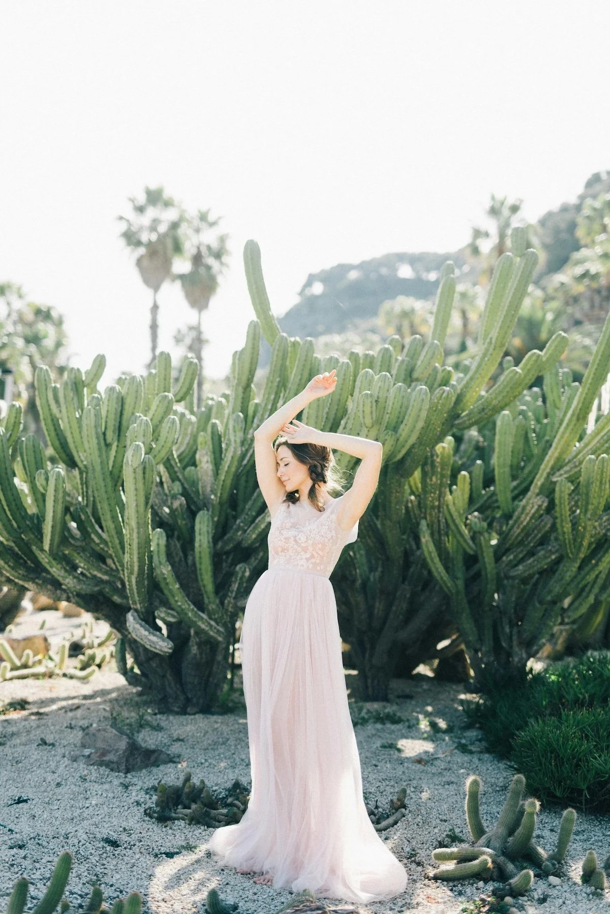 A woman in a white lace dress standing in front of a large cactus, with her arms raised and eyes closed.