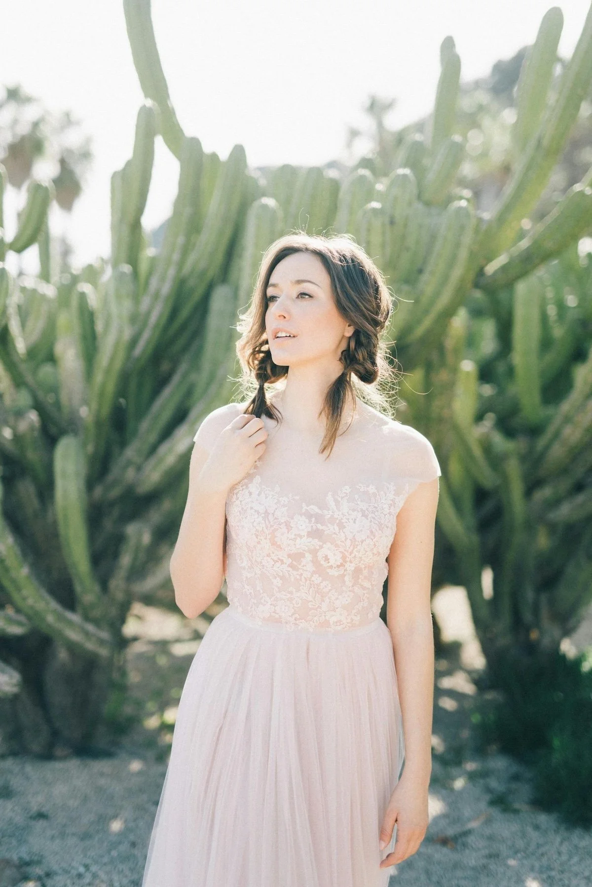 A woman with dark wavy hair standing outdoors in front of a large cactus. She is wearing a light pink, lace-top dress and holding a strand of her hair. Bright sunlight is shining down.