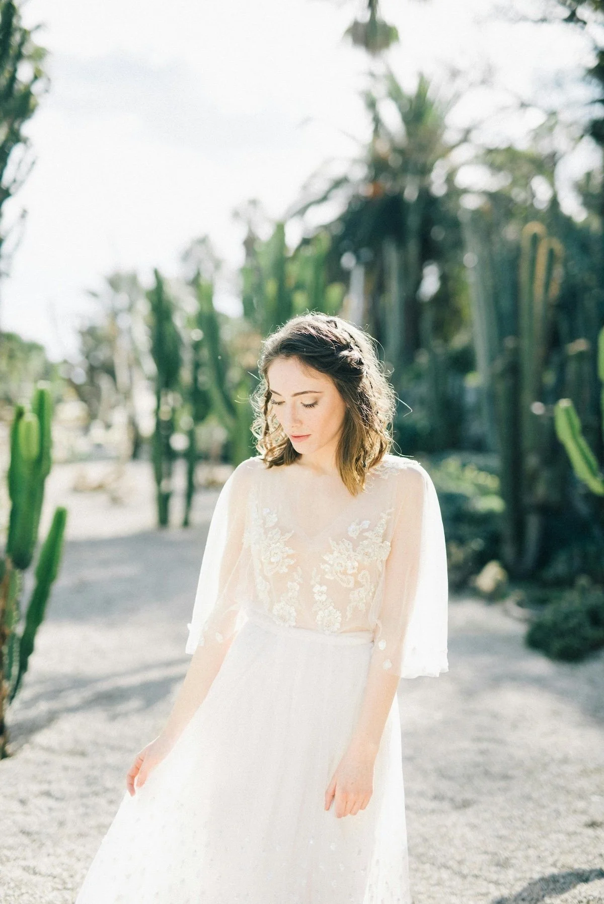 A young woman in a white lace dress with sheer sleeves, standing outdoors among tall cacti and desert plants, looking down with a serene expression.