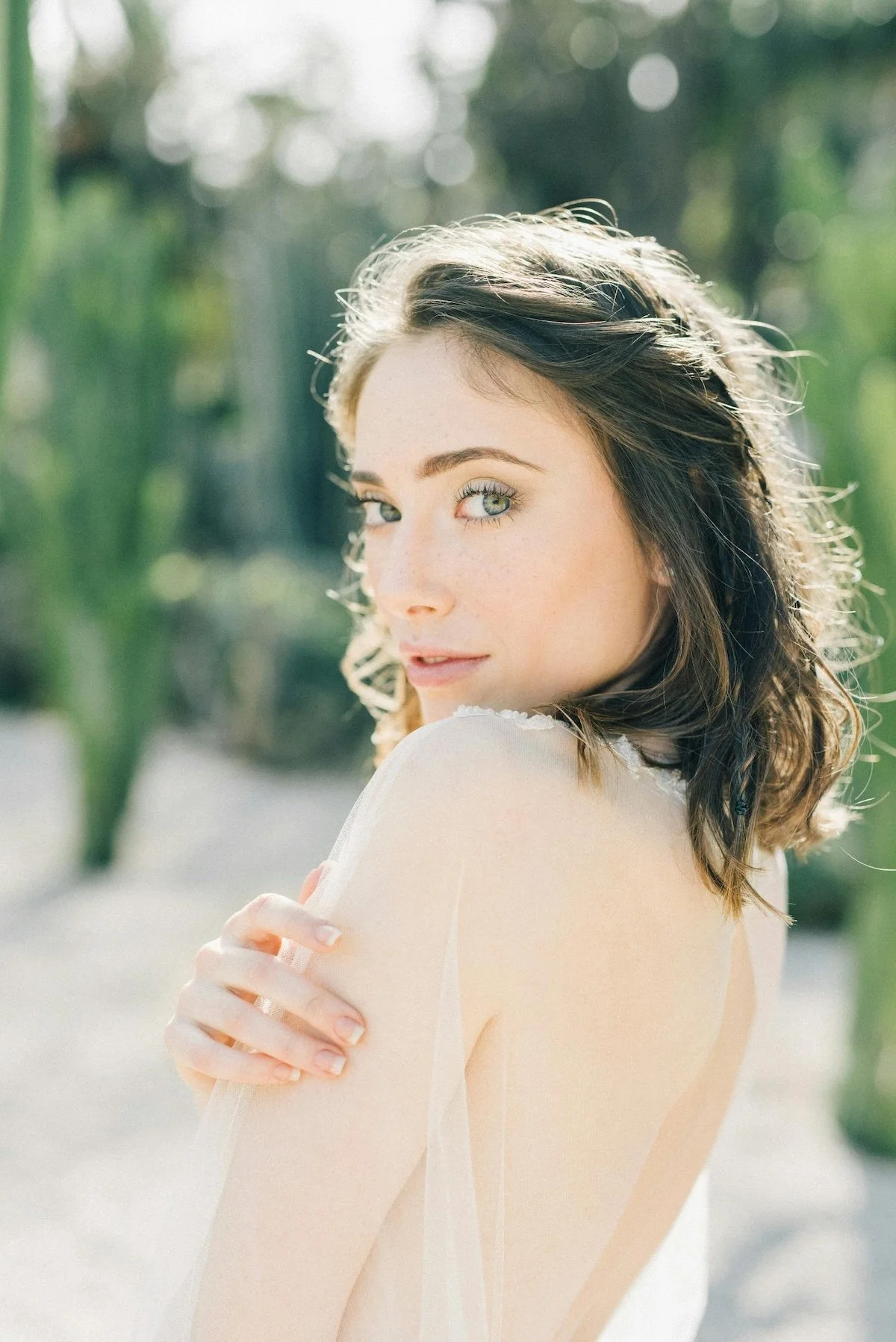 A young woman with wavy brown hair and green eyes looking over her shoulder outdoors during daytime, with sunlight highlighting her face and a blurred green background.