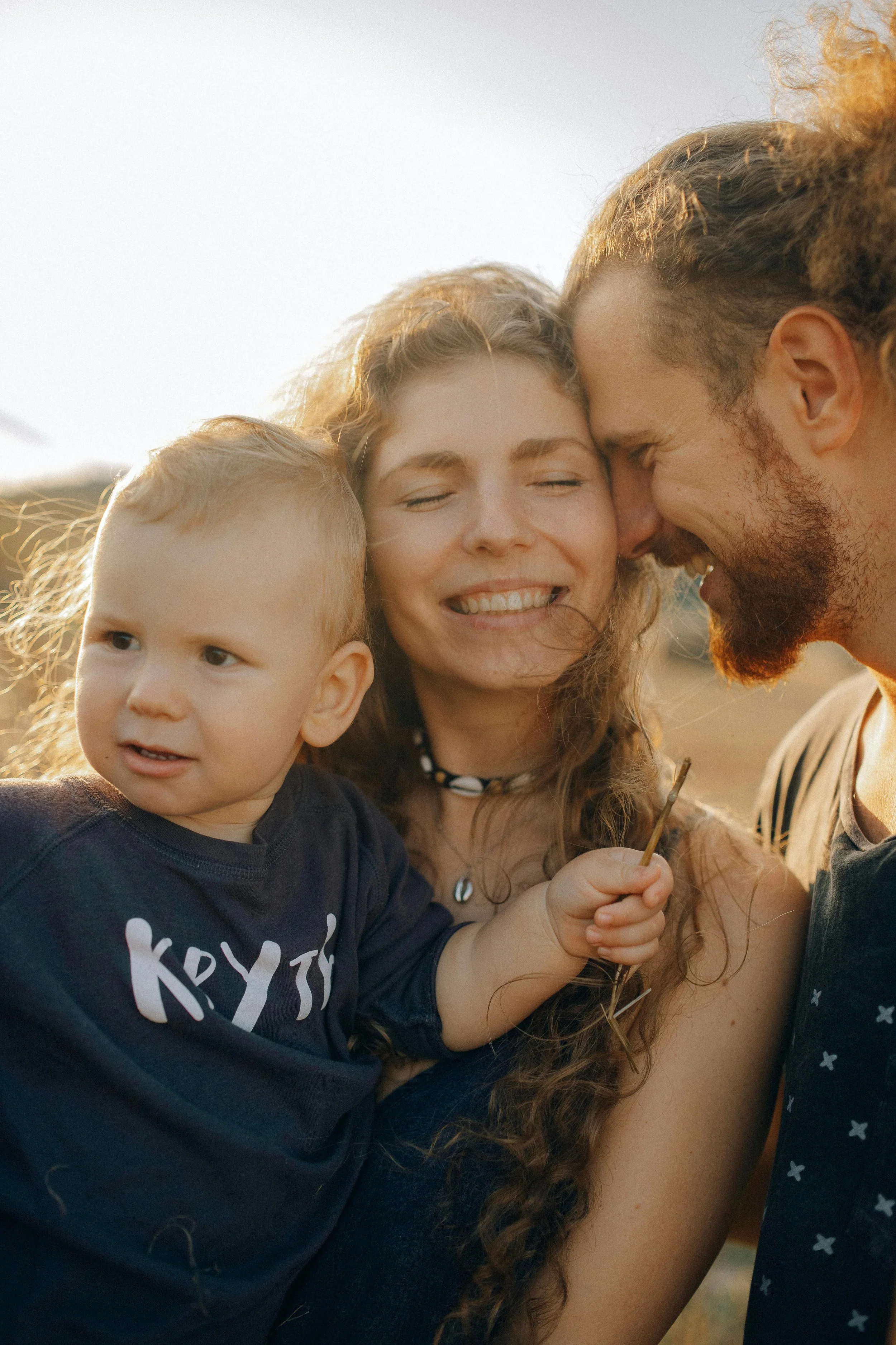 A family hugging outdoors during sunset, including a man, woman, and child, all smiling closely together.