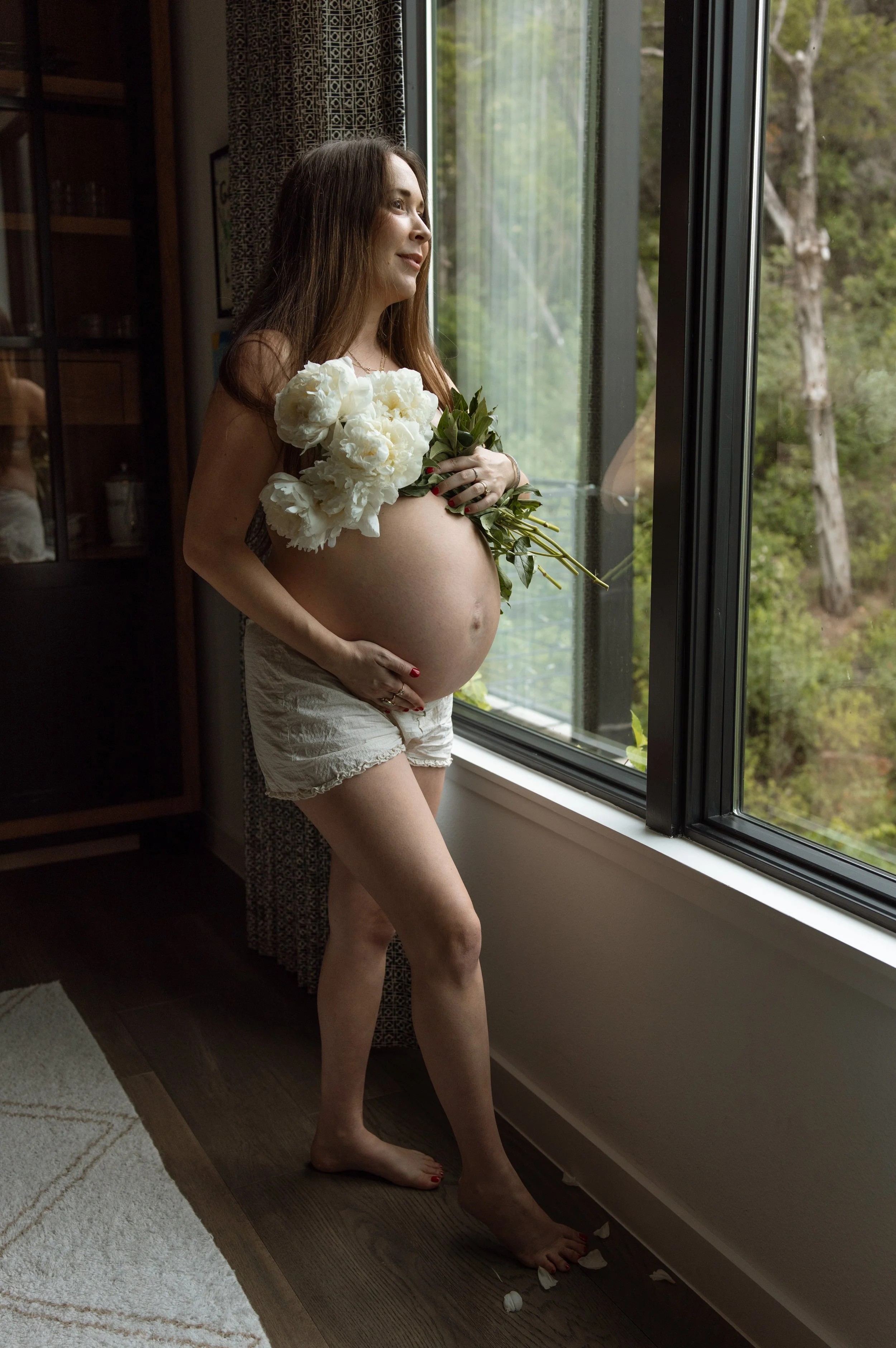 A pregnant woman standing indoors near a large window, holding a bouquet of white flowers, looking out the window with a serene expression. She is wearing light-colored shorts and has loose brown hair. Petal-like objects are scattered on the floor near her feet.