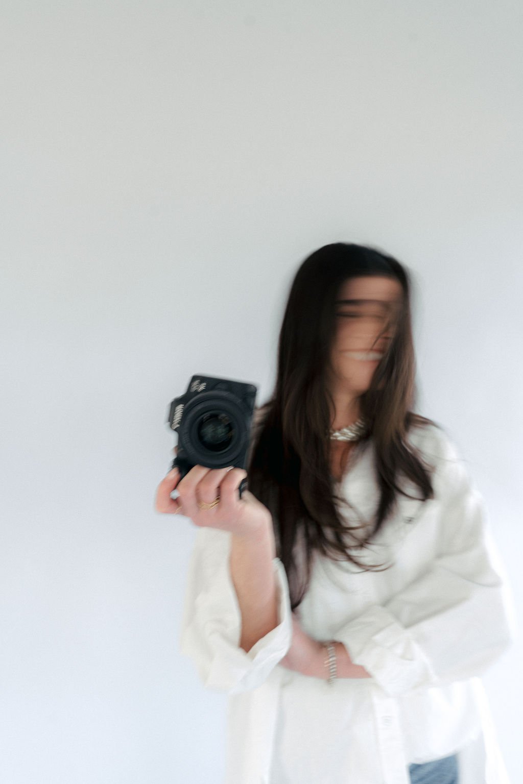 A woman with long dark hair, wearing a white shirt, is taking a selfie with a camera in her right hand, blurred face, and standing against a plain white wall.