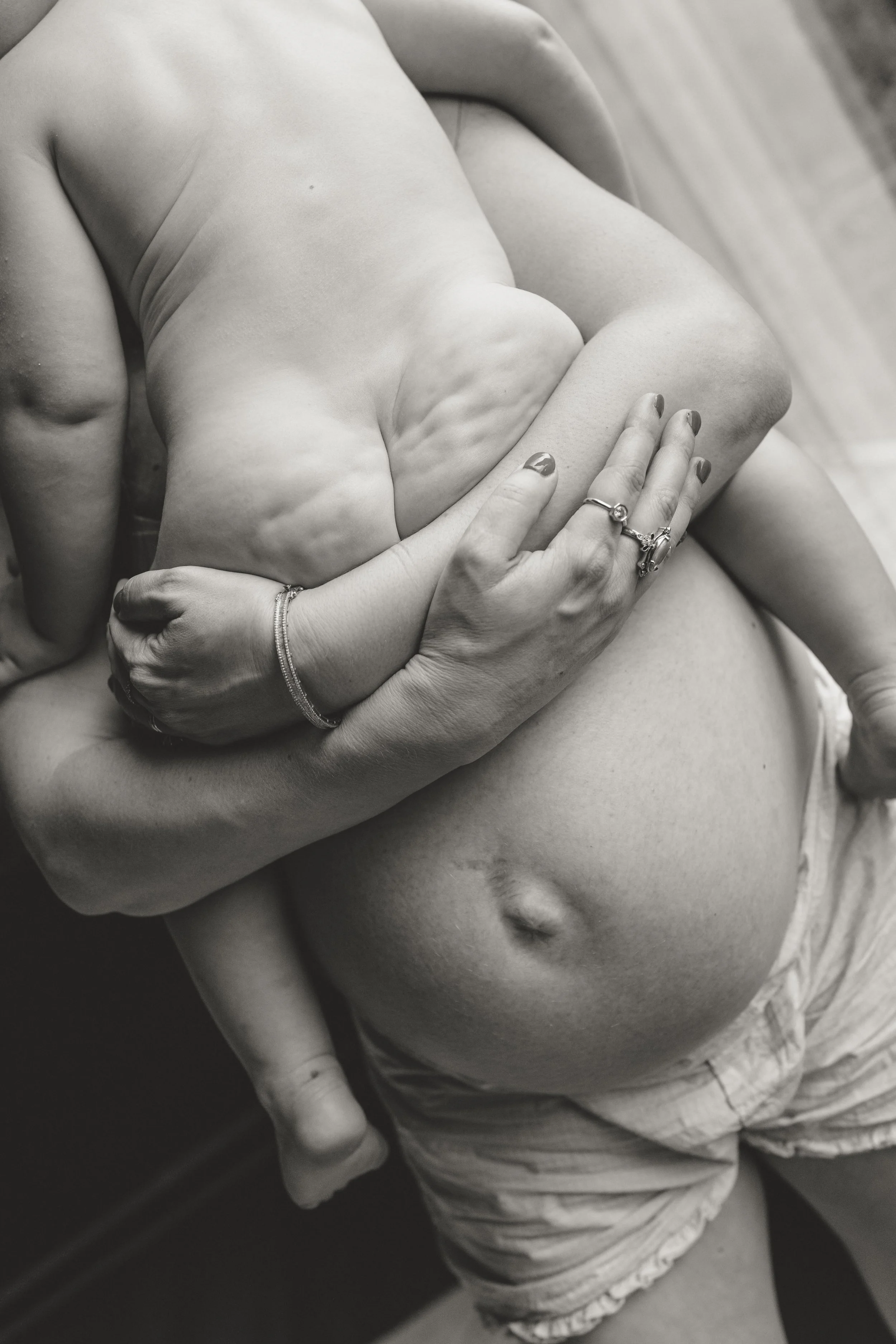 A black and white photo of a woman's torso and hand, showing her holding her pregnant belly. She is wearing rings and a bracelet, with her nails painted. The image focuses on the pregnancy and the woman's hand on her belly.