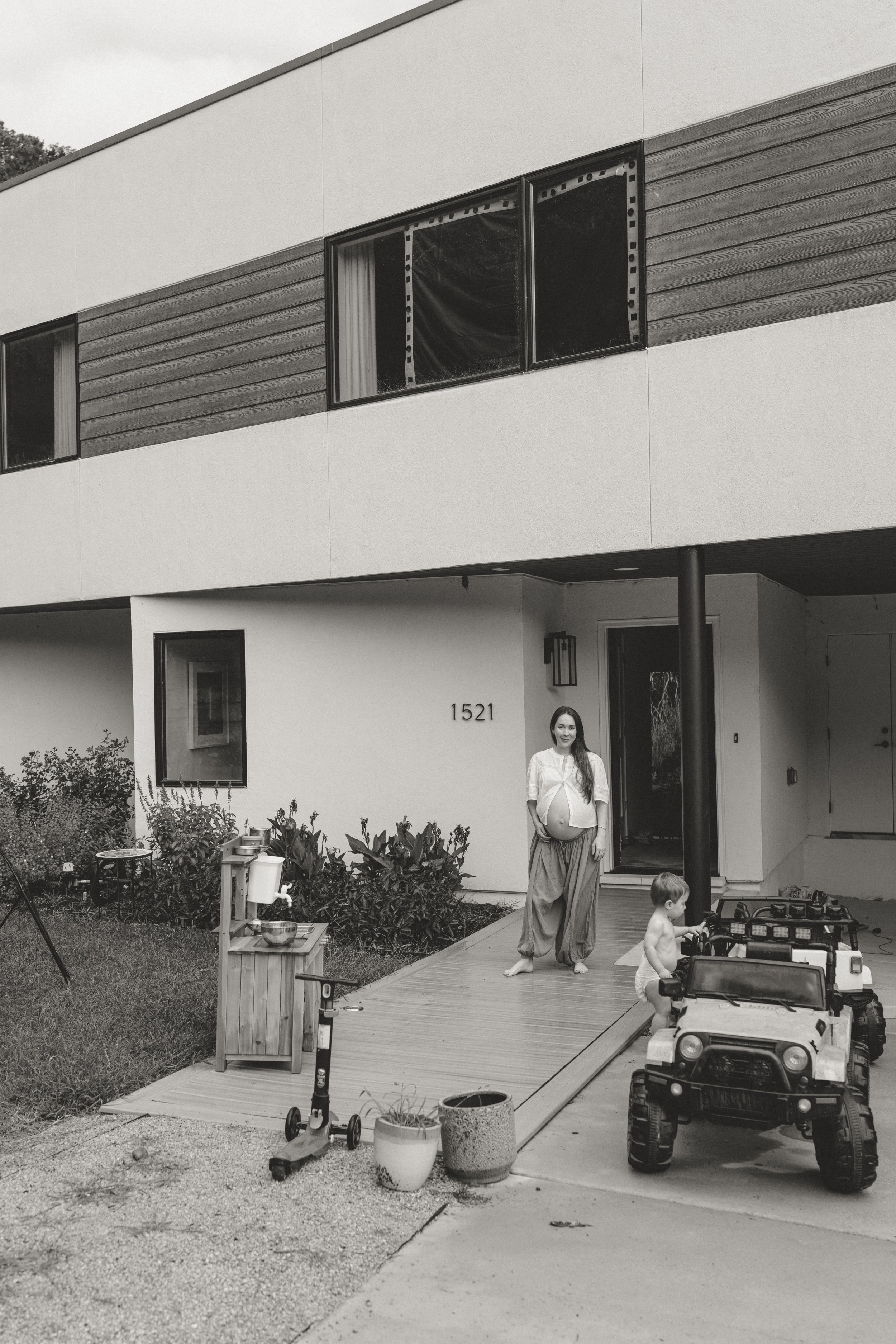A woman, whose belly appears pregnant, standing outside a modern house with a young child playing on a toy vehicle. There is a deck, potted plants, toys, and scooters in the yard. lifestyle maternity photographer in Austin 