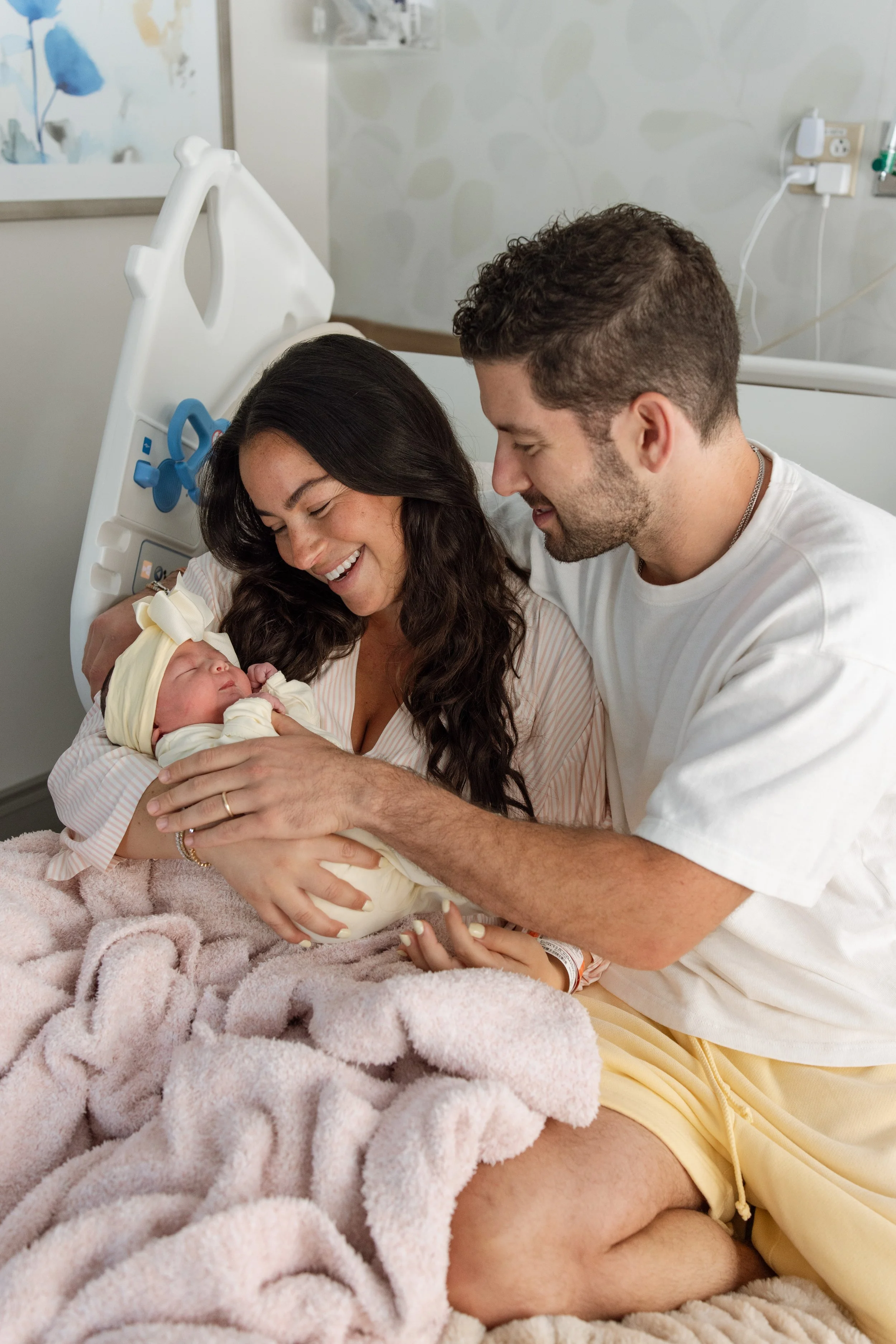 A mother and father holding their newborn baby in a hospital room, with the mother smiling and the father looking lovingly at the baby.