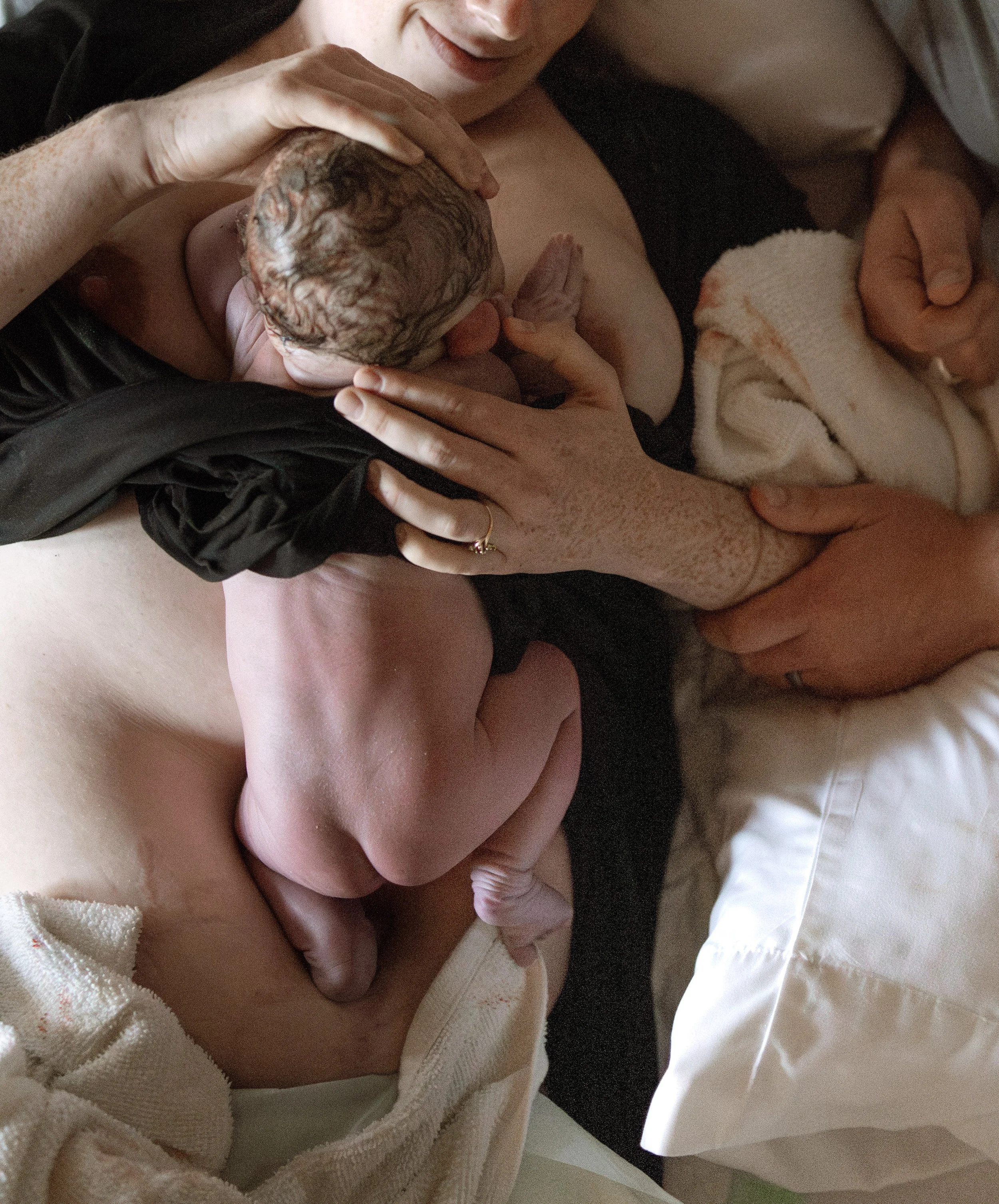 A woman holding a newborn baby, breastfeeding. The baby is wrapped in a pink blanket, and the woman wears a white sweater and black top.