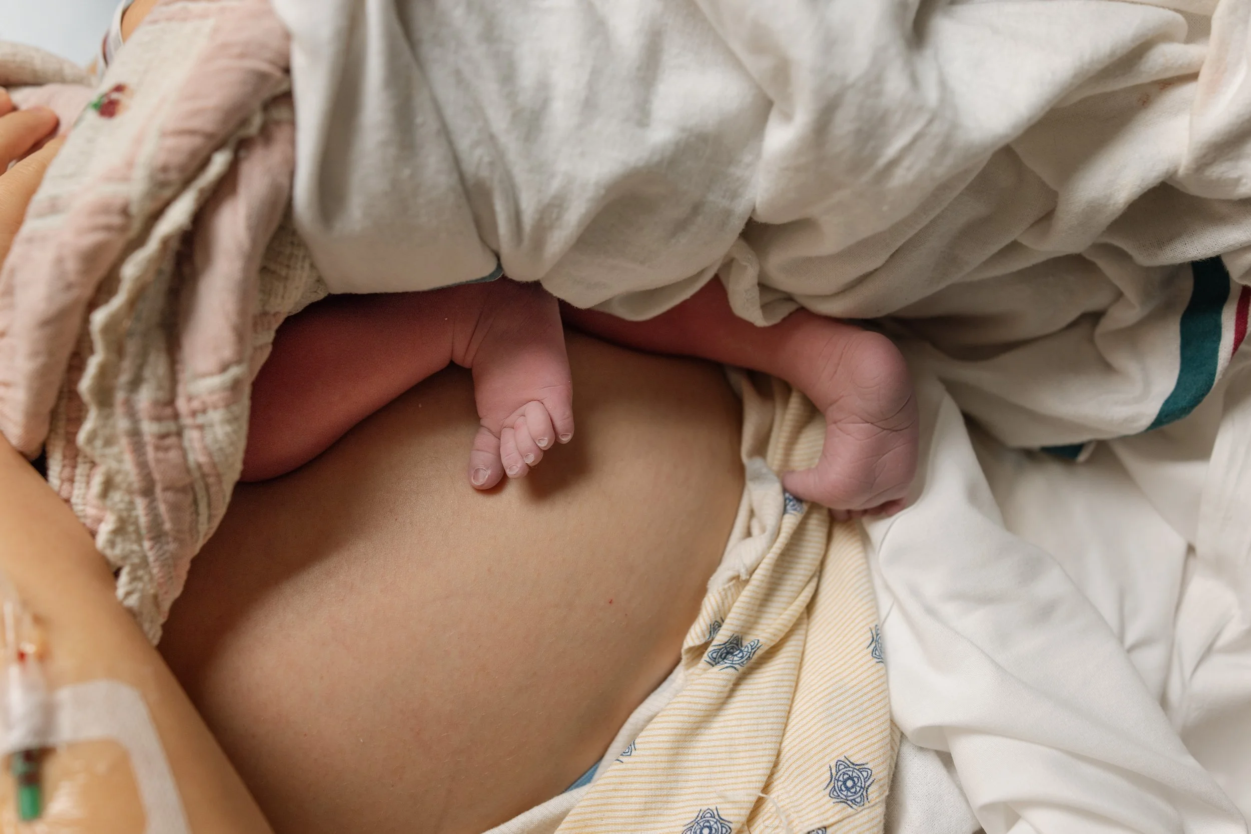 Austin birth photography photo of a newborn's feet and legs resting on her mother's stomach. they are in the hospital and have blankets on top of their bodies.