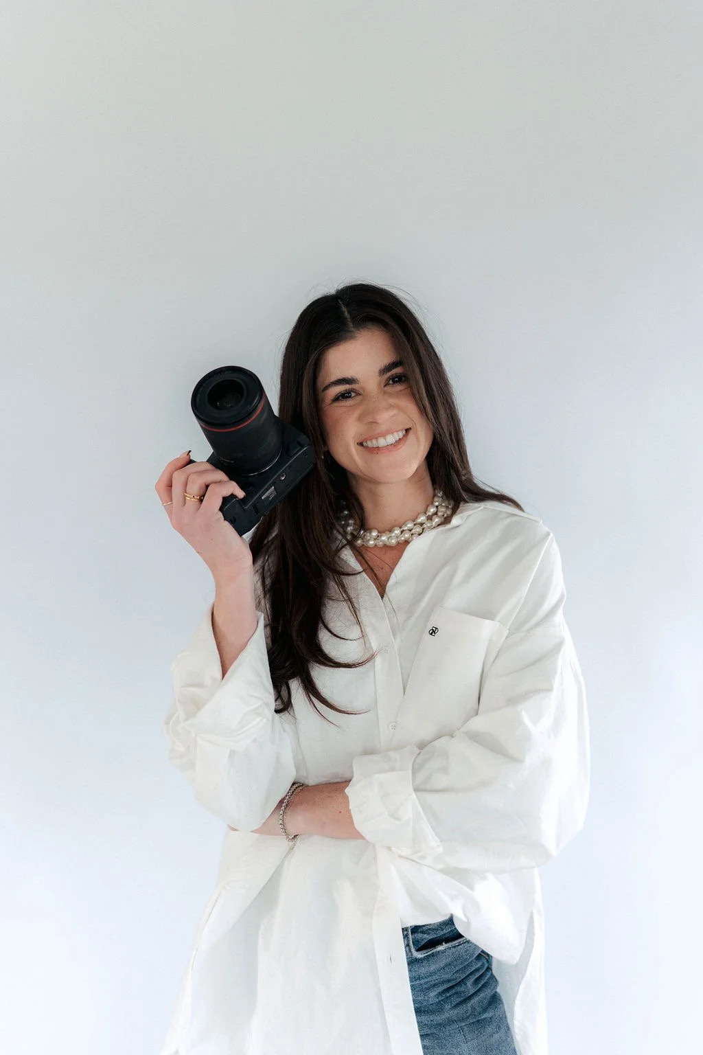 A woman with long dark hair, wearing a white shirt and pearl necklace, smiling and holding a camera to her head, standing against a plain light background.