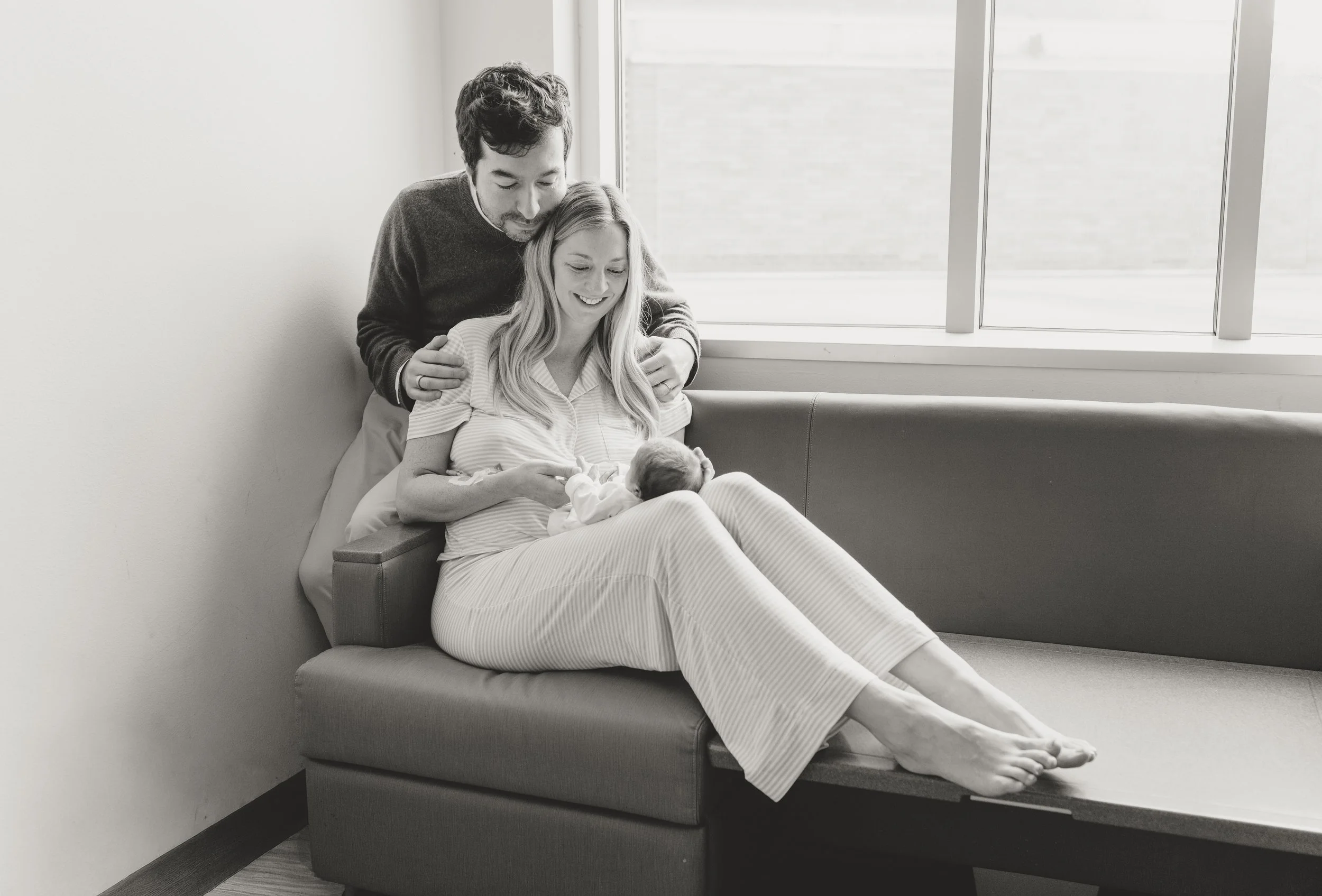 A couple with light skin sitting on a hospital bed, smiling while looking at a newborn baby wrapped in a blanket. The woman is holding the baby, and the man is leaning in affectionately. The setting appears to be a hospital room with a large window. 