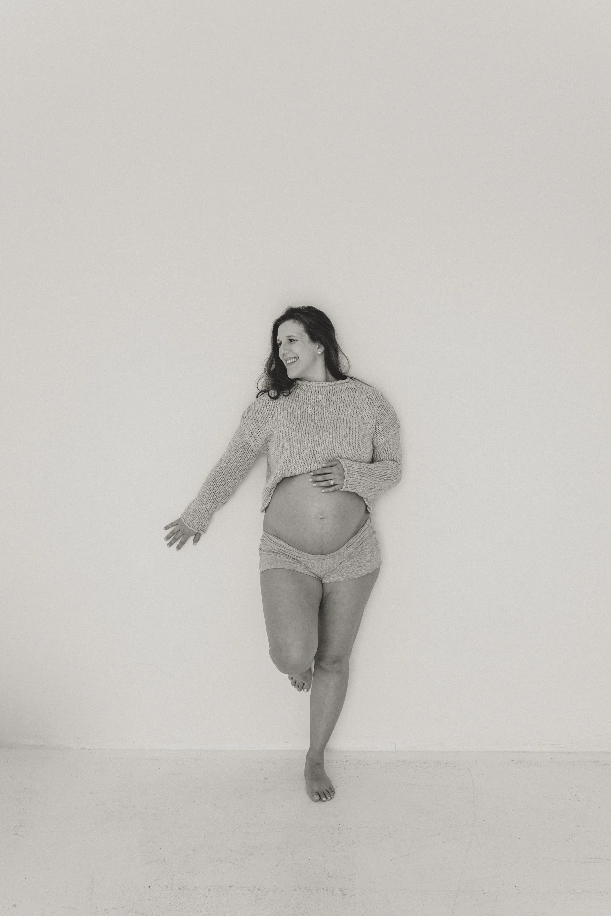A pregnant woman with dark hair smiling, wearing a sweater and shorts, standing against a plain wall, with one foot slightly raised and arms extended.