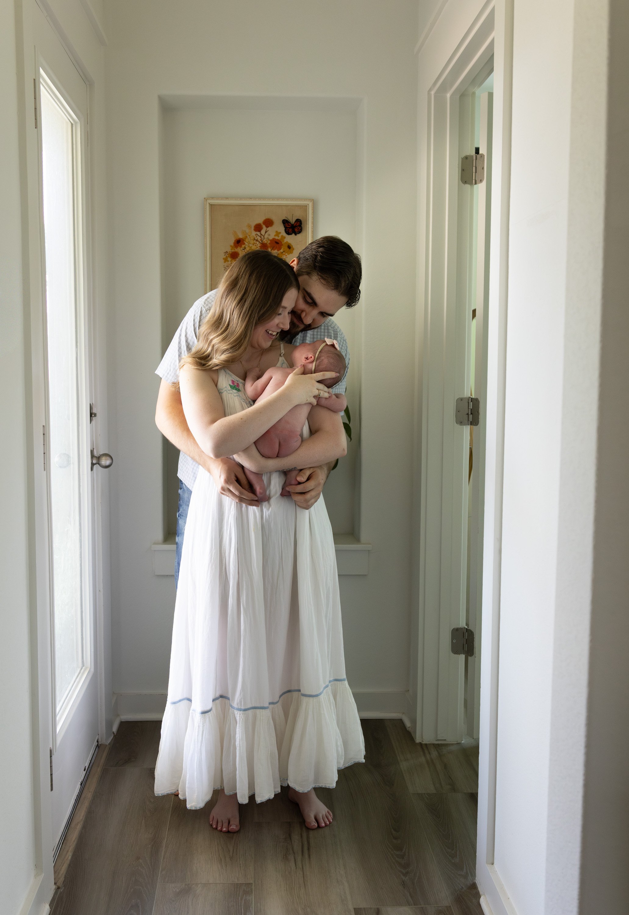 A woman and a man holding a newborn baby in a narrow hallway with white walls and wooden floors, smiling and looking at the baby. documentary newborn photography Austin Texas 