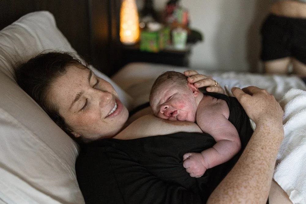 A woman lying on a bed, holding a newborn baby close to her chest, both with happy expressions. midwife assisted birth at birthing center in dripping springs Texas