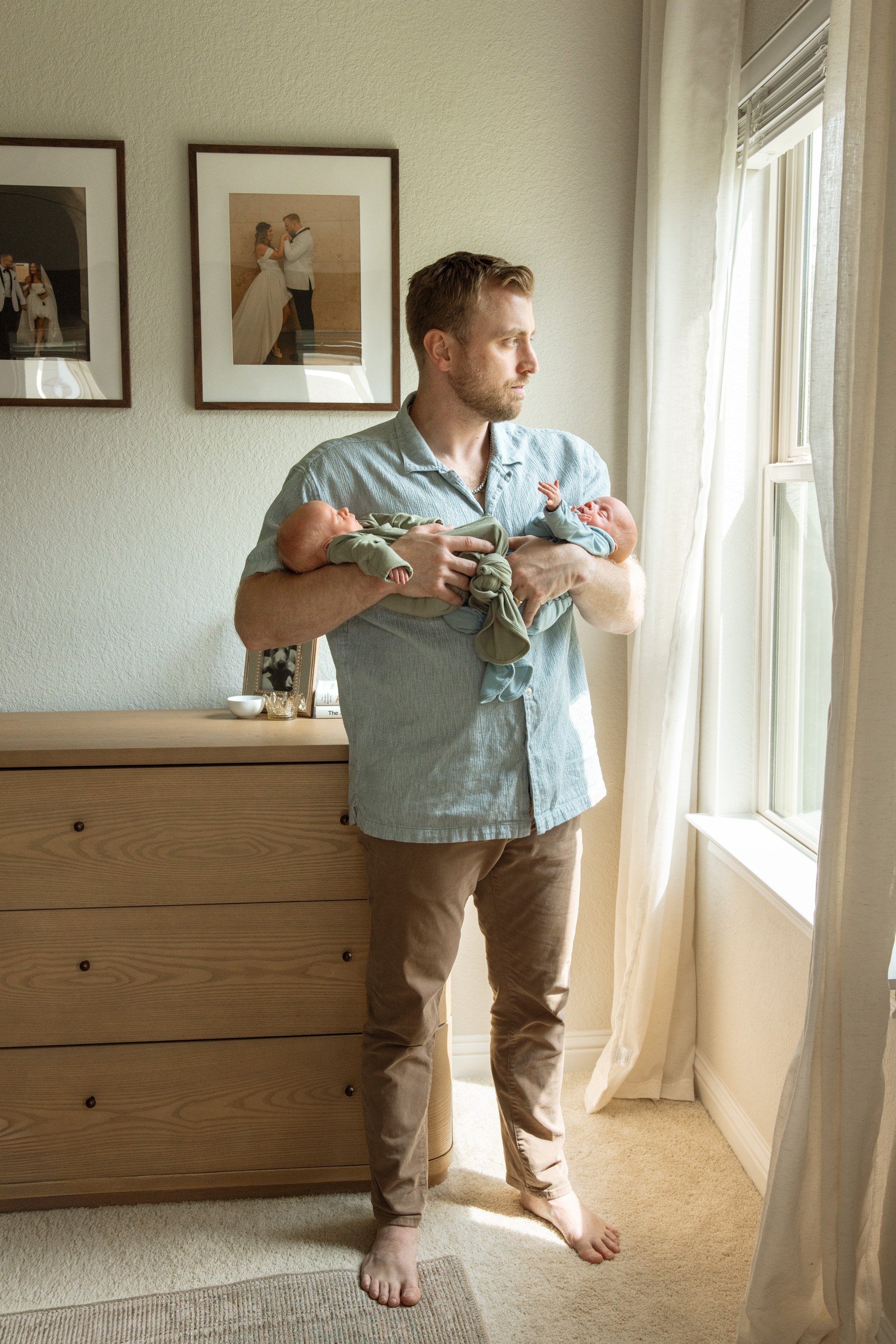 Man holding two babies by a window in a bedroom with framed wedding photos on the wall. Austin Texas in home newborn photography 