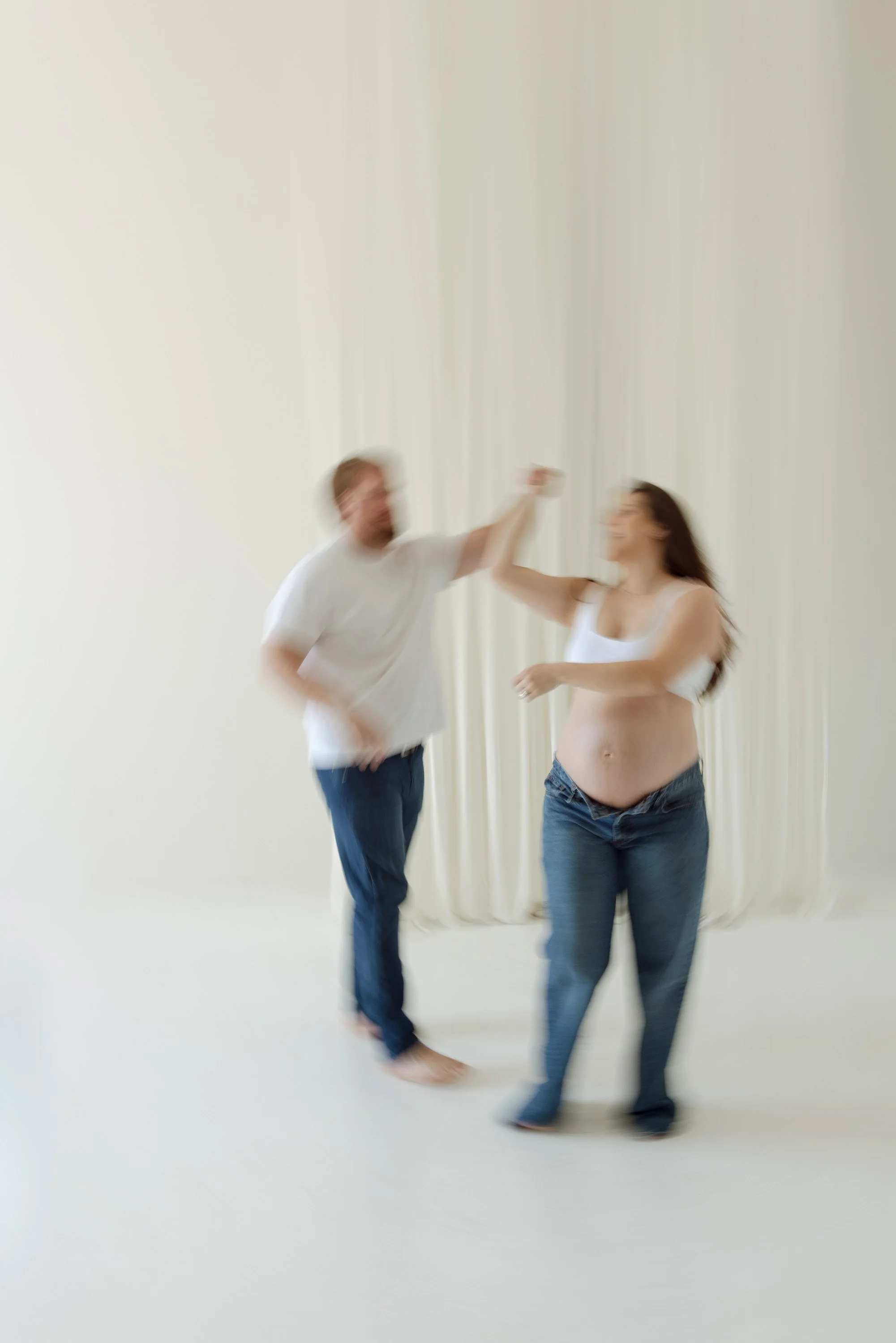 A blurry photo of a man and a pregnant woman dancing together in front of a white curtain. The woman has long hair and is wearing a white tank top and jeans, while the man has short hair and a beard, wearing a white T-shirt and jeans. editorial studi