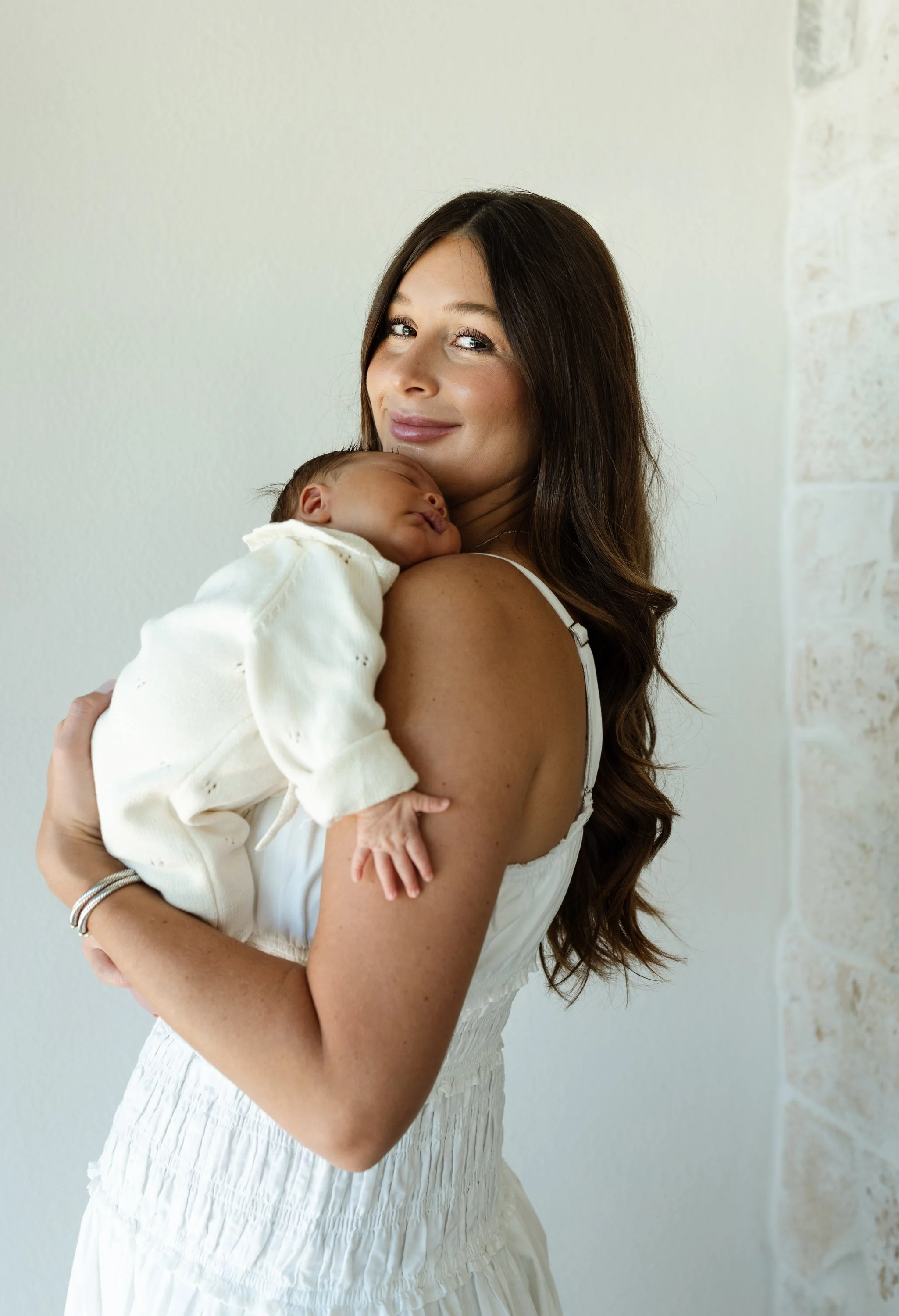 Austin first time mom is holding her newborn daughter and wearing a white dress while smiling for newborn photos.