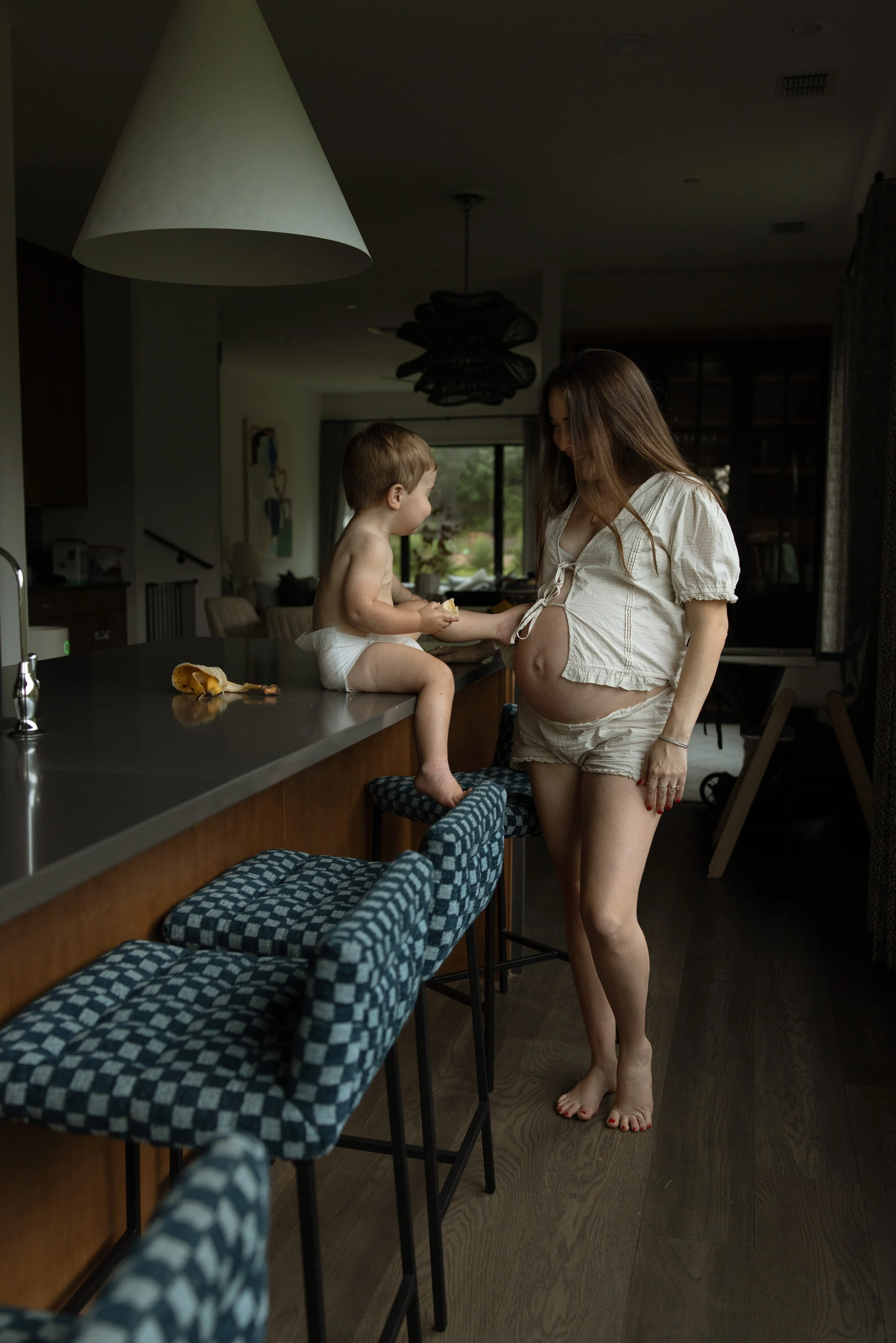 A pregnant woman with long hair wearing a light-colored top and shorts stands barefoot in a kitchen, looking at a young boy sitting on the kitchen counter. The boy, shirtless and in a diaper, is interacting with the woman. The kitchen has a dark coun