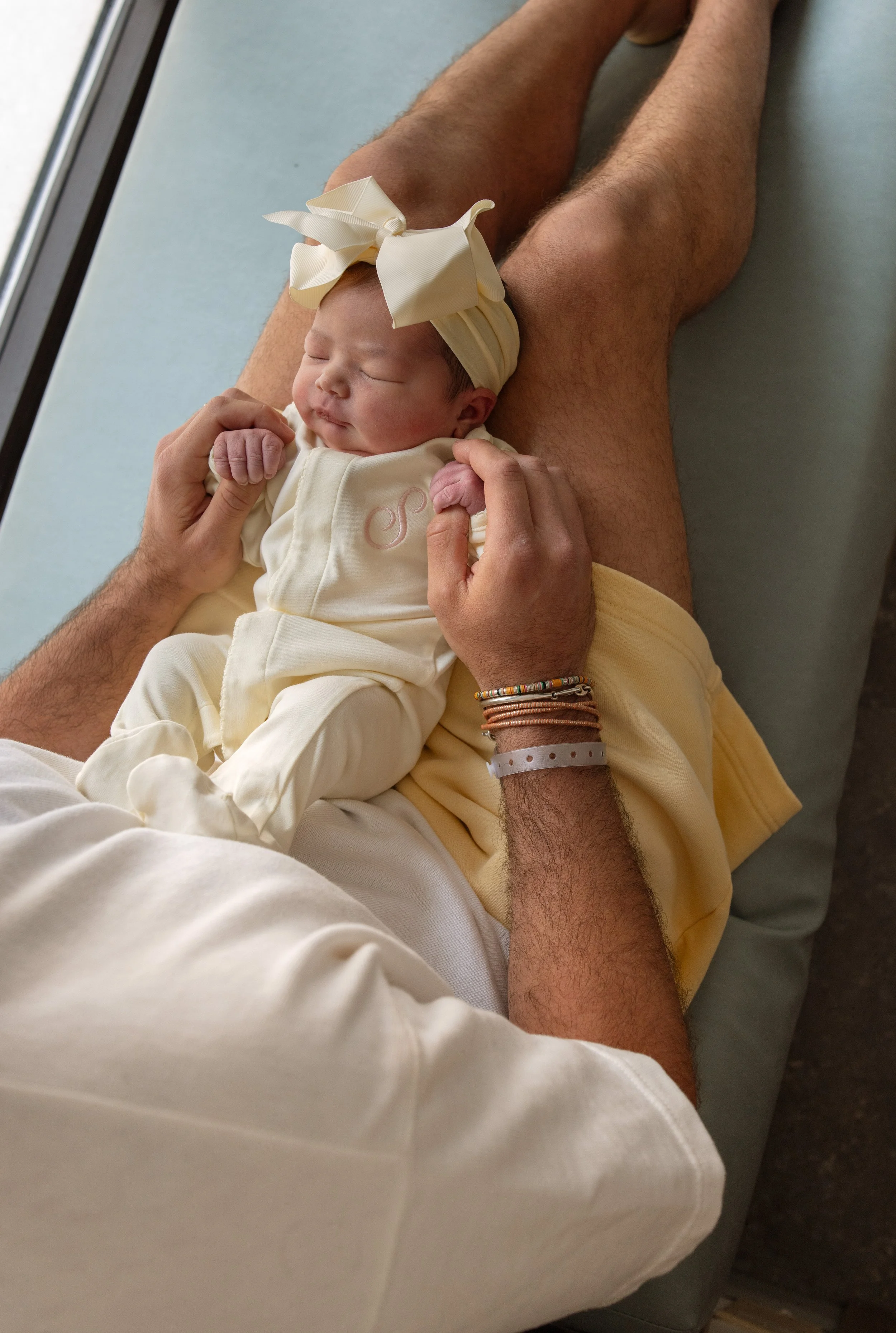 A newborn baby with a large cream-colored bow headband, sleeping peacefully on an adult's lap in a hospital or clinic setting. fresh 48 photos Austin Texas