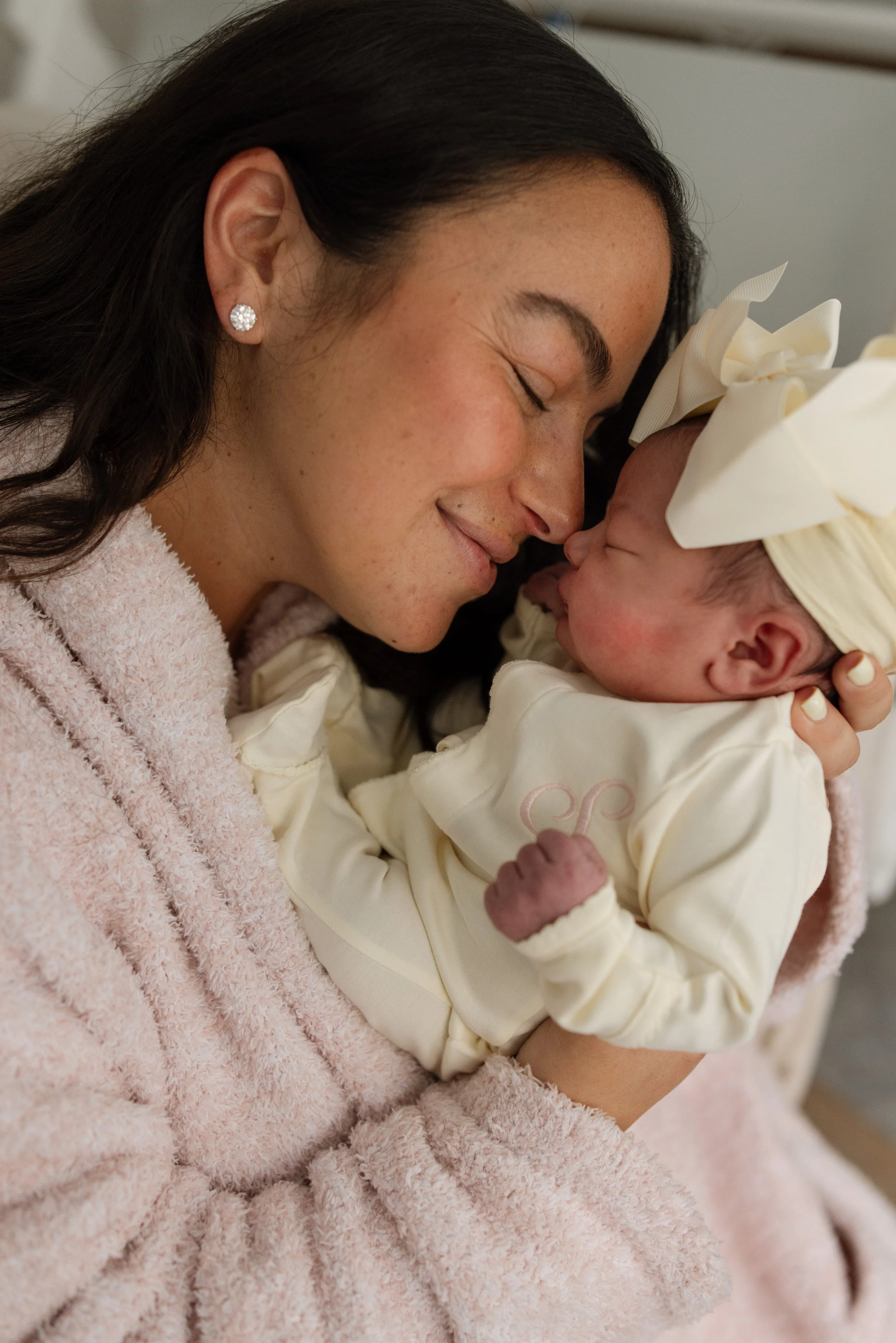 A woman with dark hair and pearl earrings tenderly holds a newborn baby dressed in cream-colored clothing and a matching headband, with their foreheads touching in a close, loving moment. fresh 48 photoshoot Austin Texas
