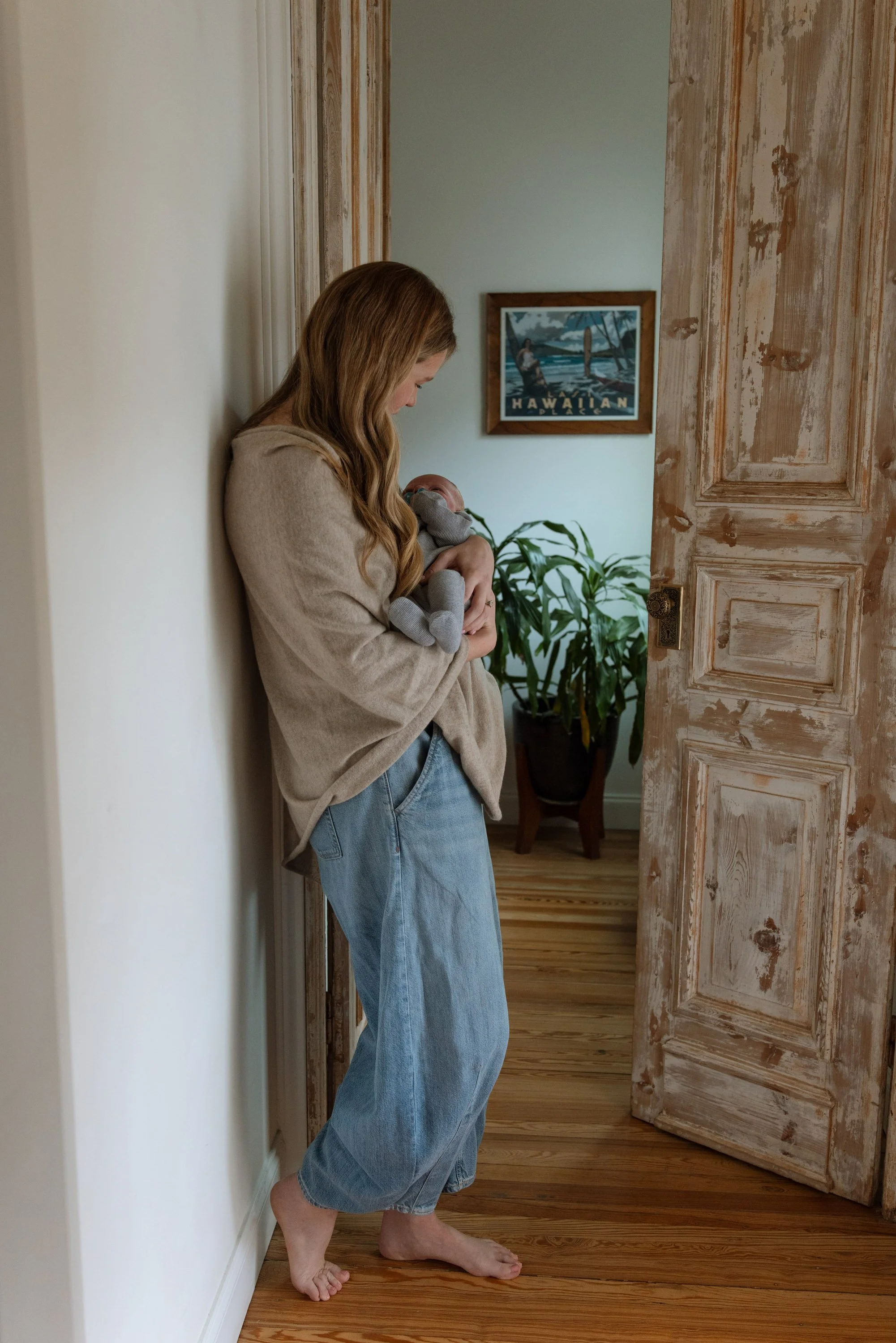 A woman with long wavy hair, wearing a beige sweater and light blue jeans, is standing barefoot in a room, holding a baby wrapped in a gray blanket. The woman is leaning against a white wall, looking down at the baby. The room has wooden flooring, a plant, and a framed picture of a Hawaiian scene on the wall in the background. The door is partially open, revealing the interior of the room.
