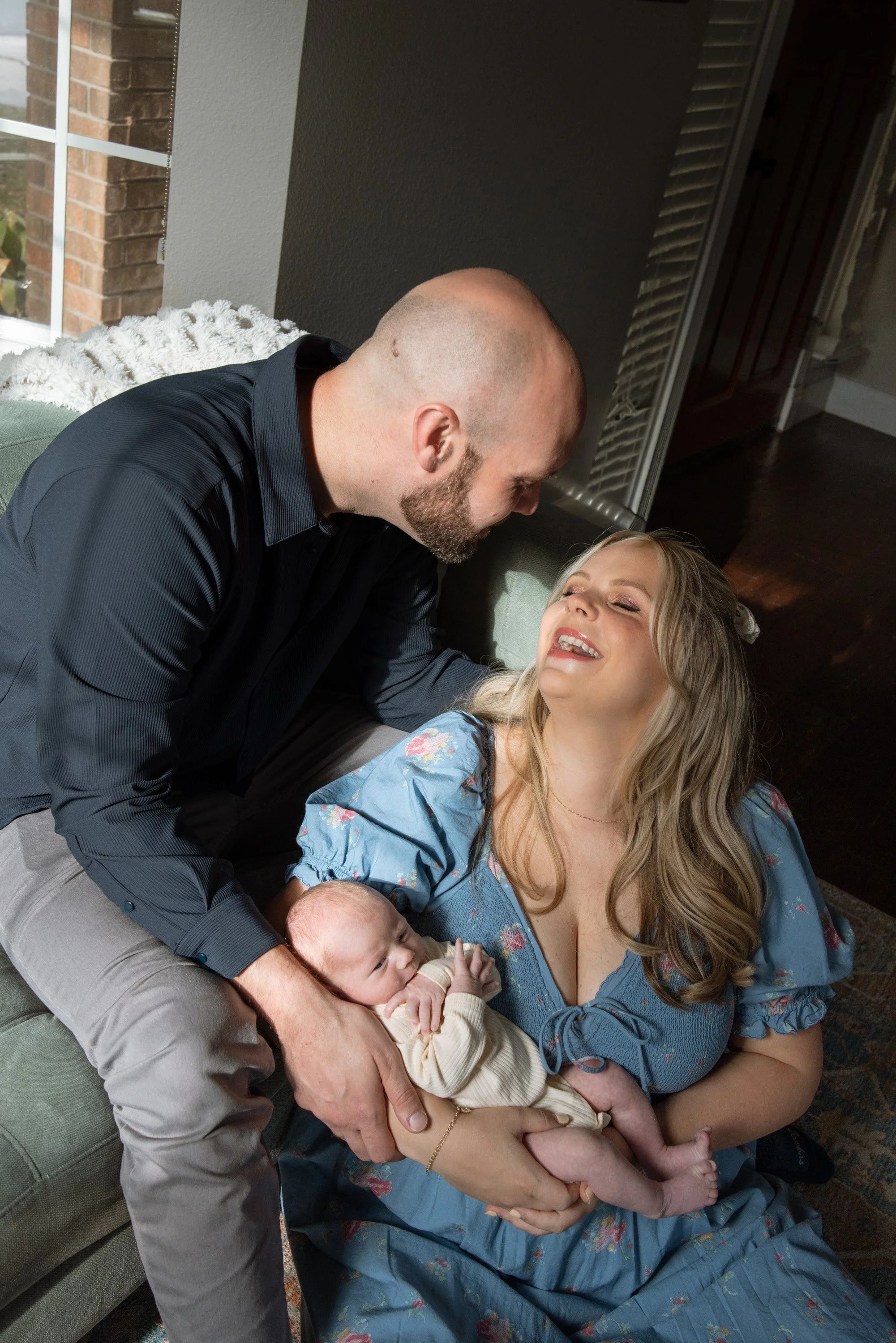A man and woman share a joyful moment with a baby on a couch in a living room. The man is leaning over the woman, smiling, while she is lying back, also smiling. The woman holds a small baby, who is looking up at her. newborn photographer in dripping